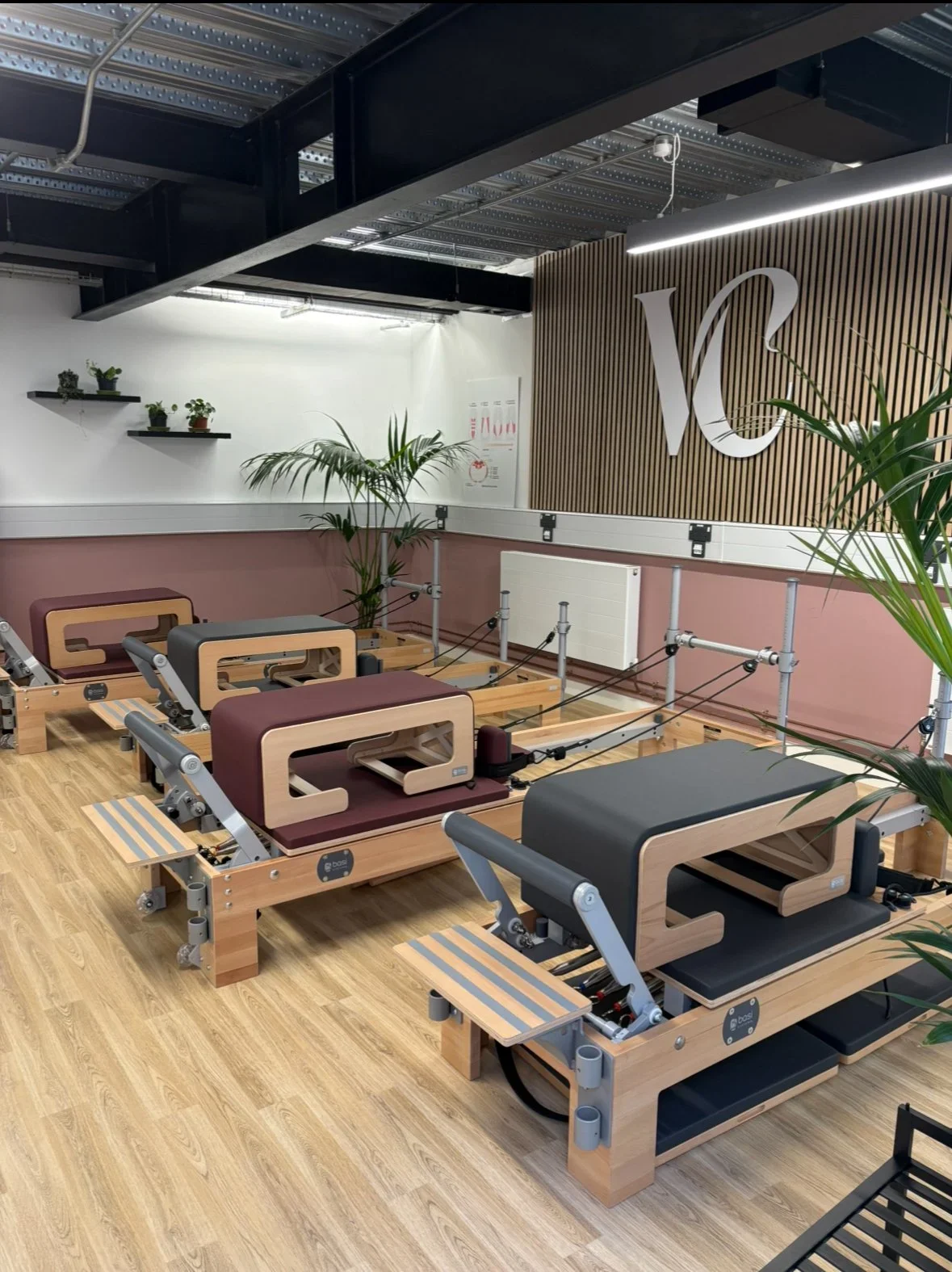 Indoor Pilates studio with wooden reformer machines, green plants, and a large logo on a wooden wall.