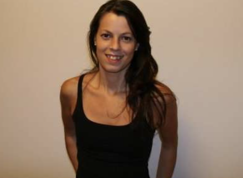 A woman with long dark hair wearing a black sleeveless top, standing against a plain beige wall, smiling at the camera.
