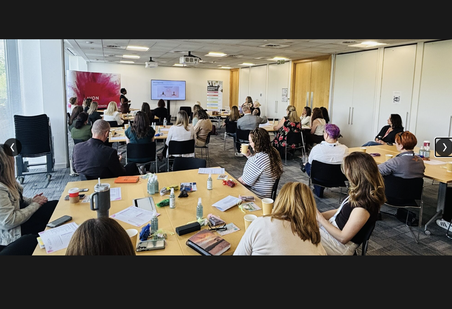 A conference room filled with people attending a presentation. The presenter stands near a screen with a slide, and there is a large AVON banner on the left side of the room. Attendees sit at tables with papers, drinks, and personal items.