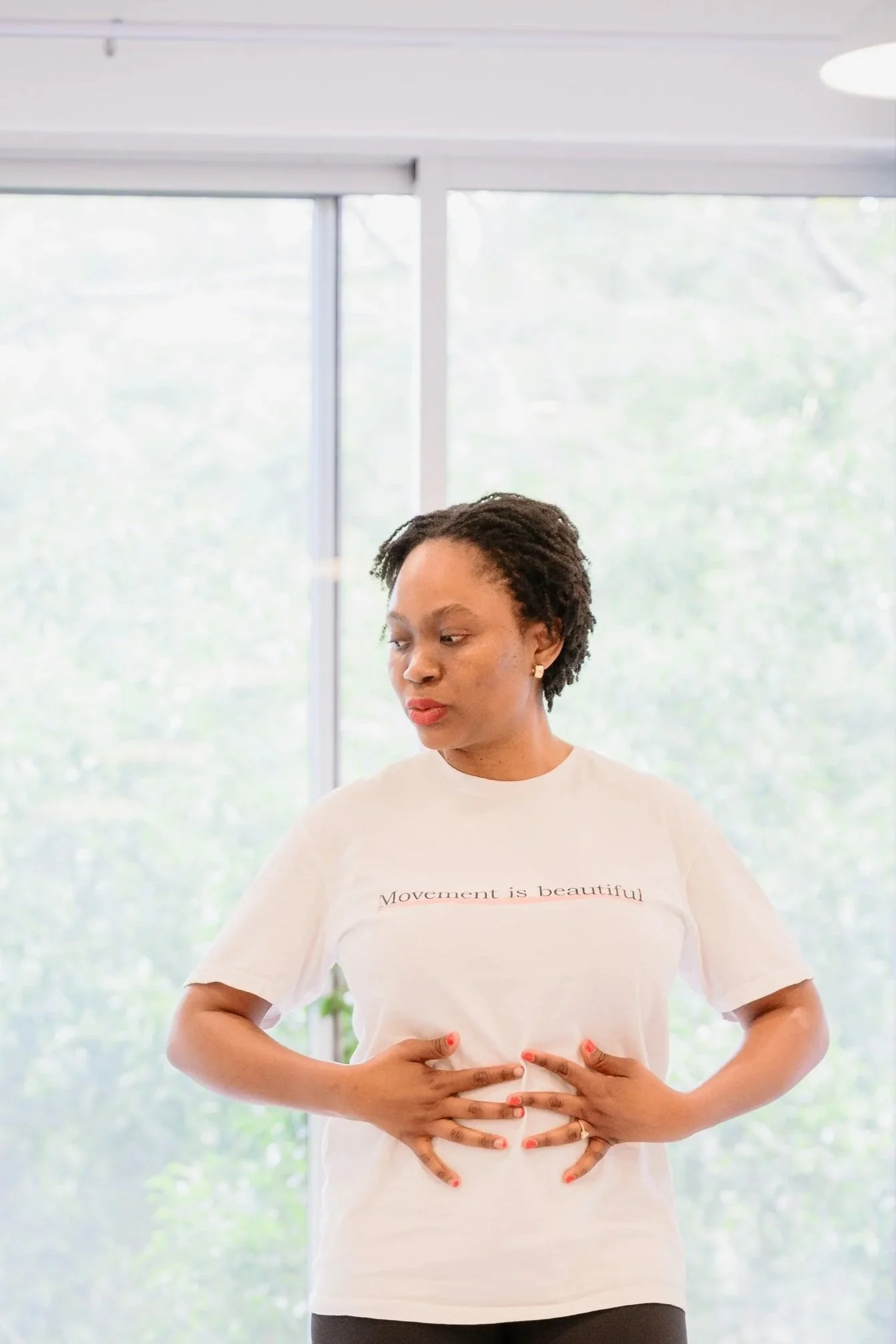 A woman standing indoors with large windows behind her, wearing a white t-shirt that reads 'Movement is beautiful'. She has short curly hair, red lipstick, and is holding her stomach with both hands.