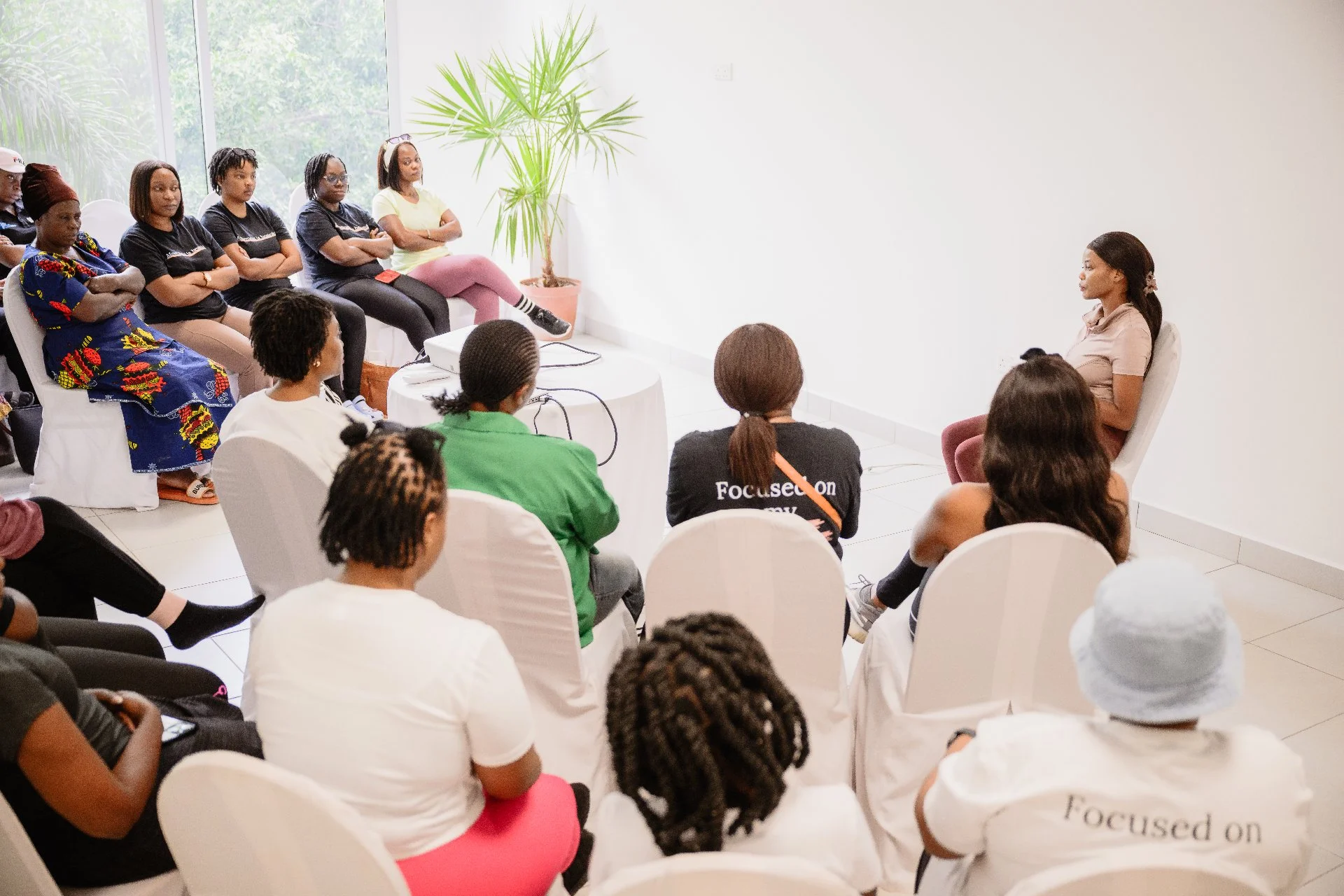 A woman is speaking to a group of women seated in a bright, minimalistic room with white walls and a large window. The audience appears attentive, sitting in white chairs, with some wearing black shirts with white text.