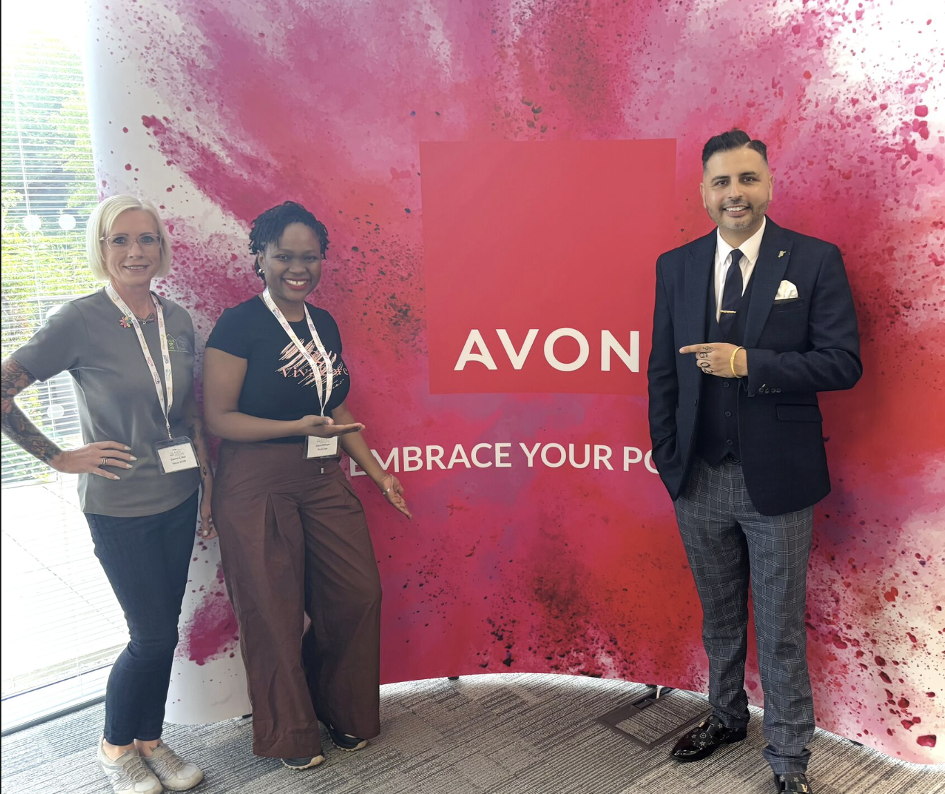 Three people standing in front of a pink and red background with the AVON logo and the slogan "Embrace Your Power." They are at a professional event, smiling and posing for the photo.