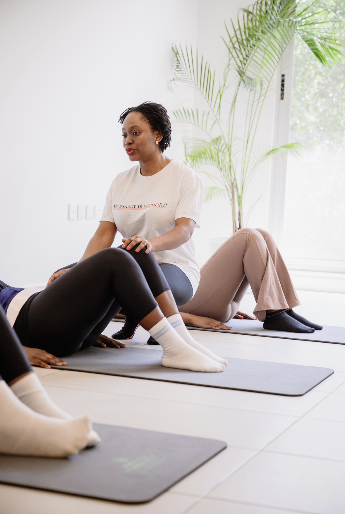 A woman instructing a fitness class in a bright room with large windows and a tall plant in the background, wearing a white t-shirt and black leggings.