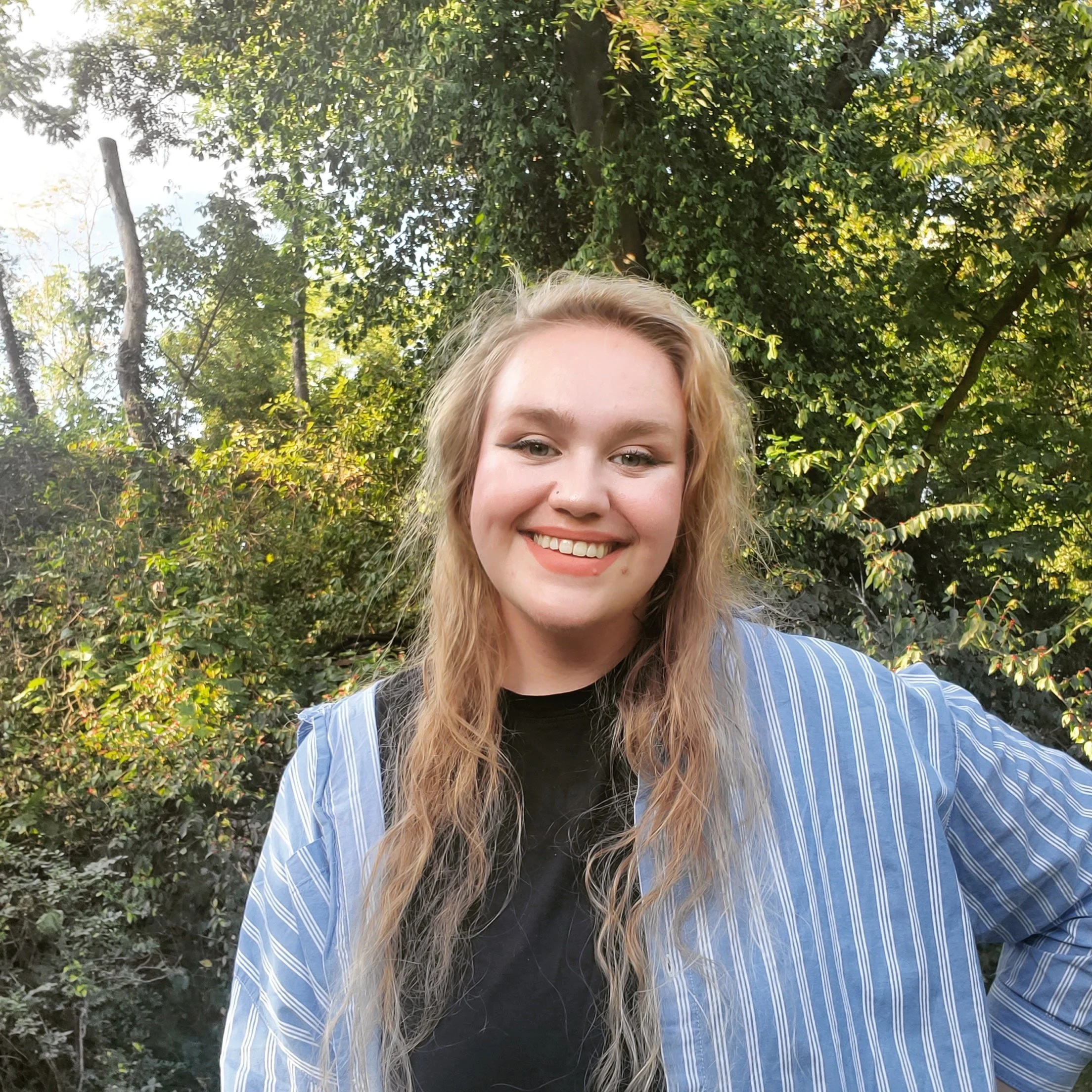 Close-up of a young woman with blonde hair smiling outdoors in front of a background of green trees.