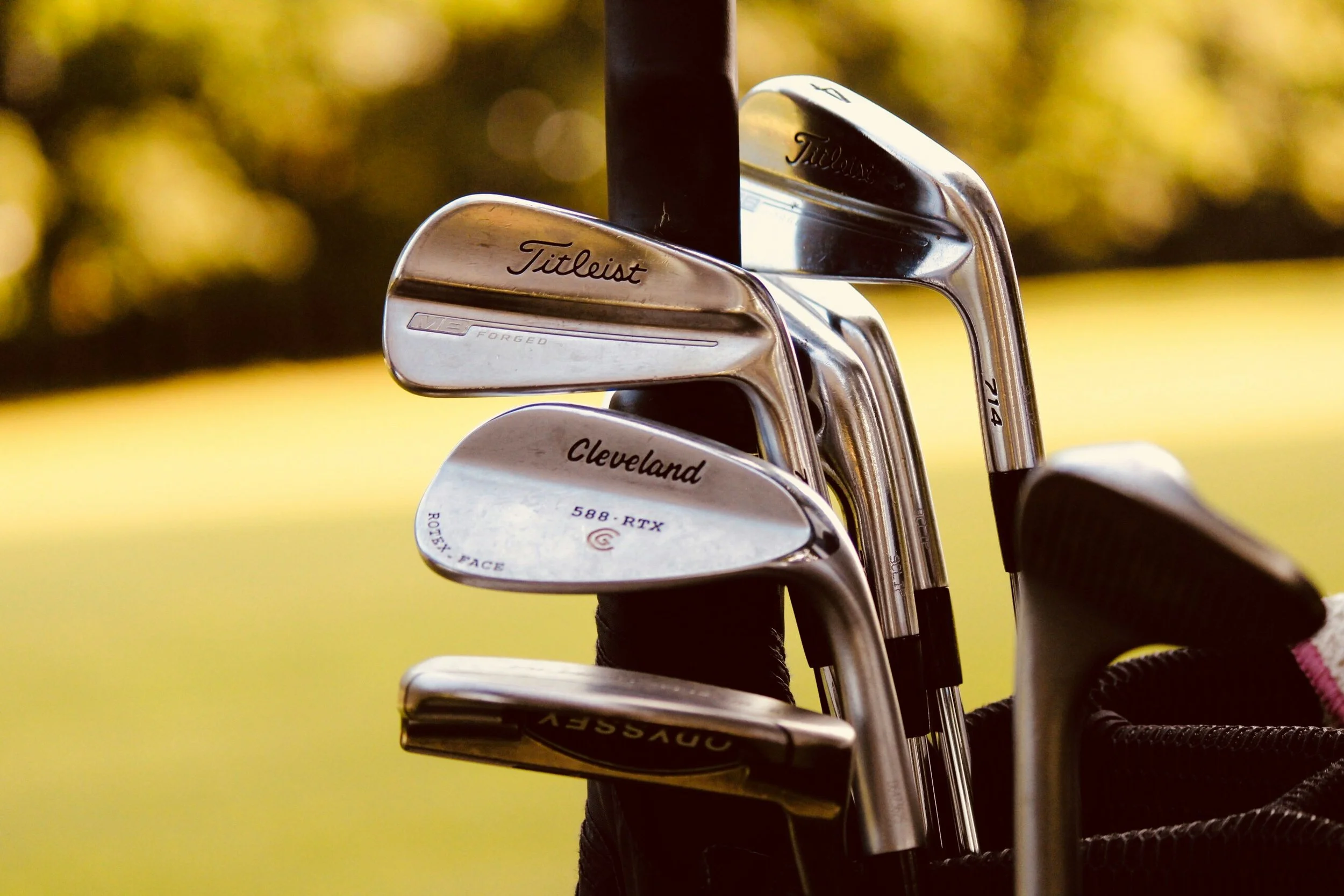 Set of golf clubs in a golf bag, with focus on the club heads displaying brand names like Titleist and Cleveland, against a blurred natural background.