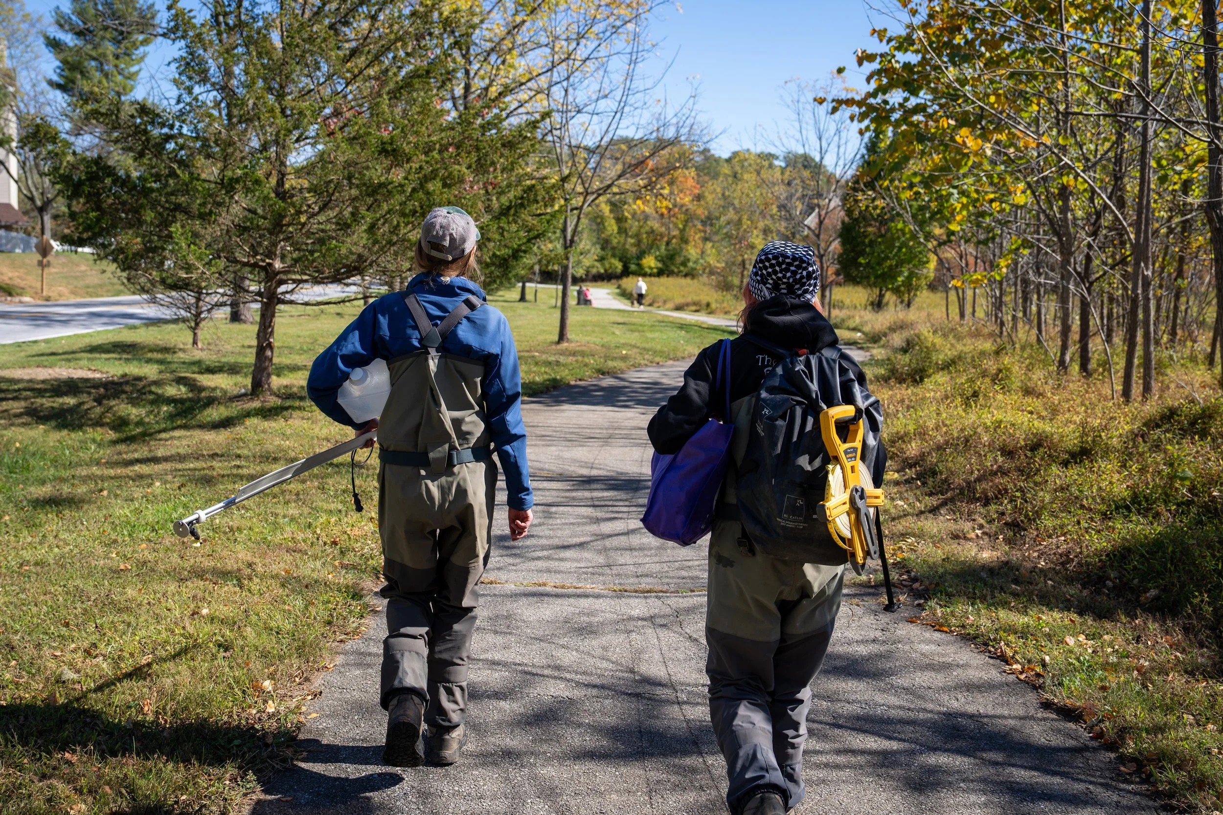 Two people walking on a paved trail through a park or nature area with trees, some with fall foliage, on a sunny day.