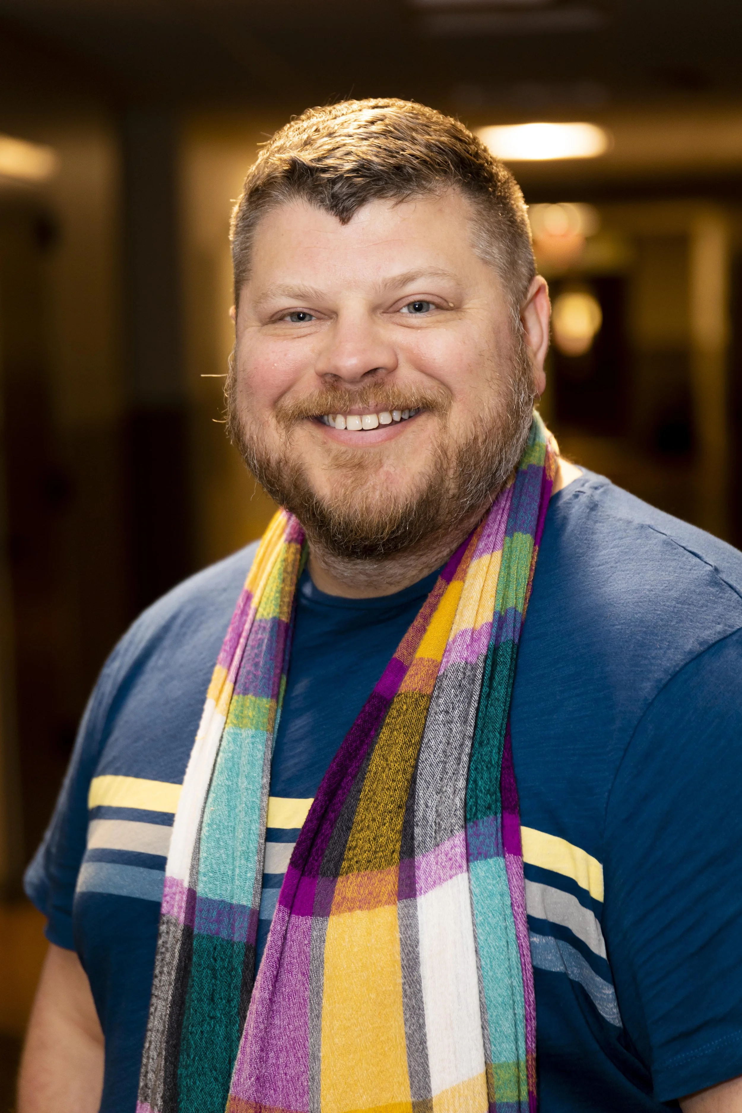 A smiling man with a beard and short hair wearing a blue shirt and a colorful plaid scarf. The background is a warmly lit indoor setting.