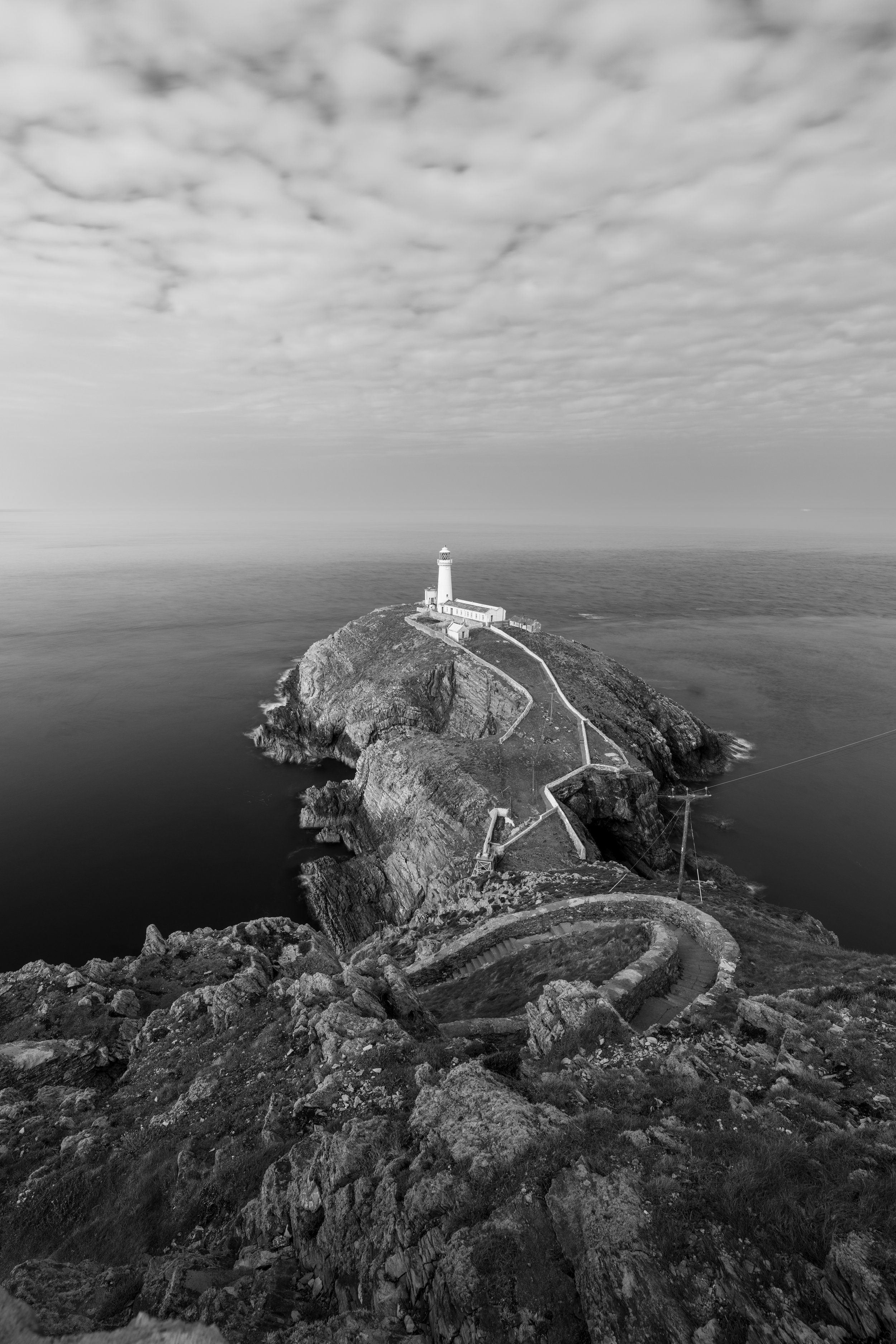 A black and white photo of a lighthouse on a rocky island, with stairs leading up to it, surrounded by water and a cloudy sky.