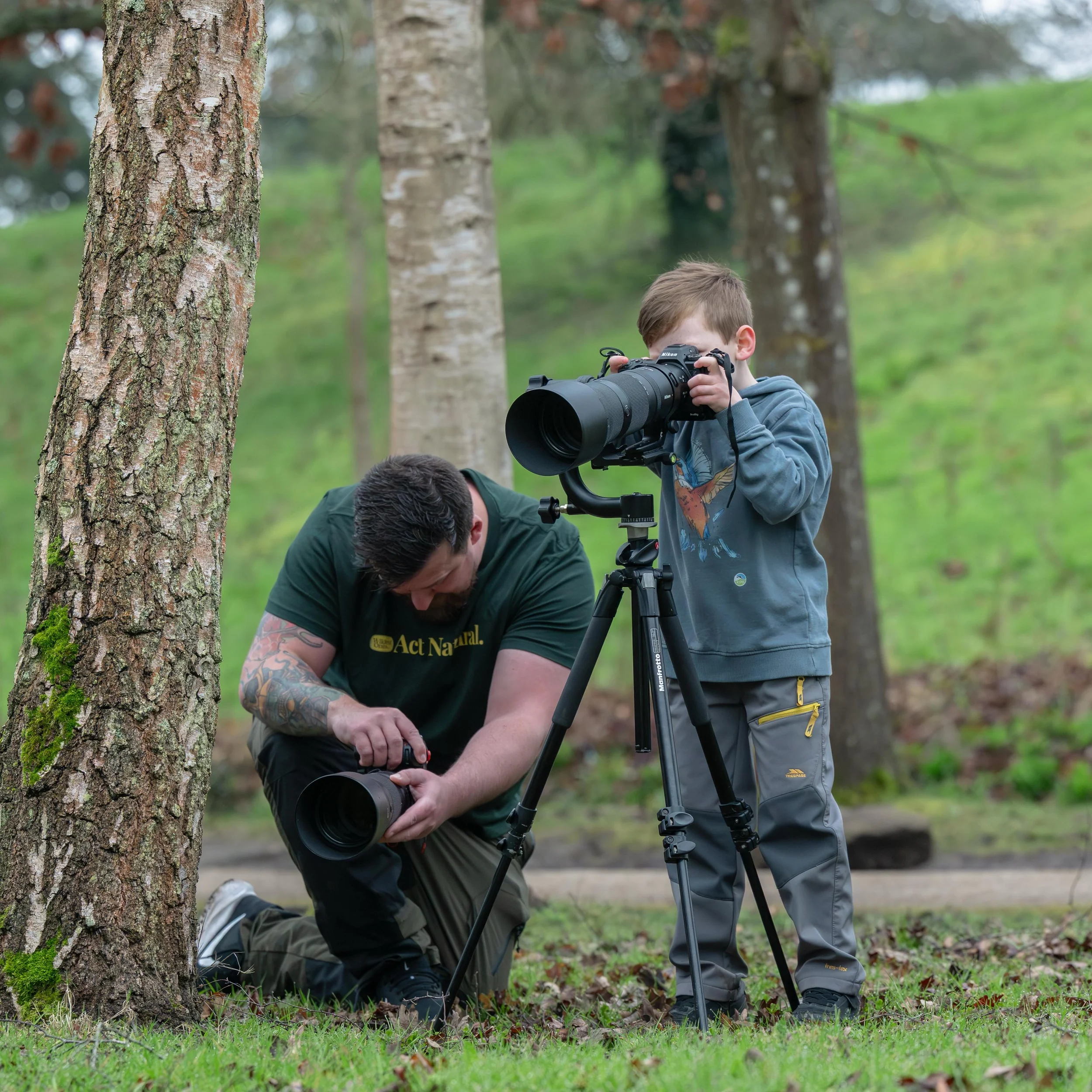 A man kneeling next to a young boy, both focused on setting up a camera on a tripod in a wooded outdoor area.