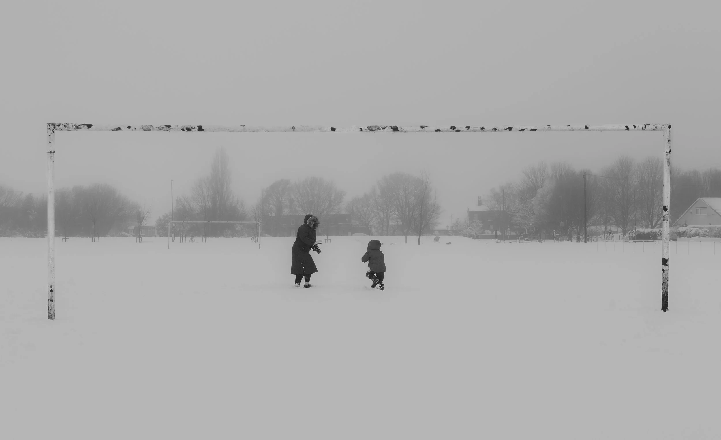 A snowy soccer field with a parent and child playing under a goalpost, with trees and houses in the background, in black and white.