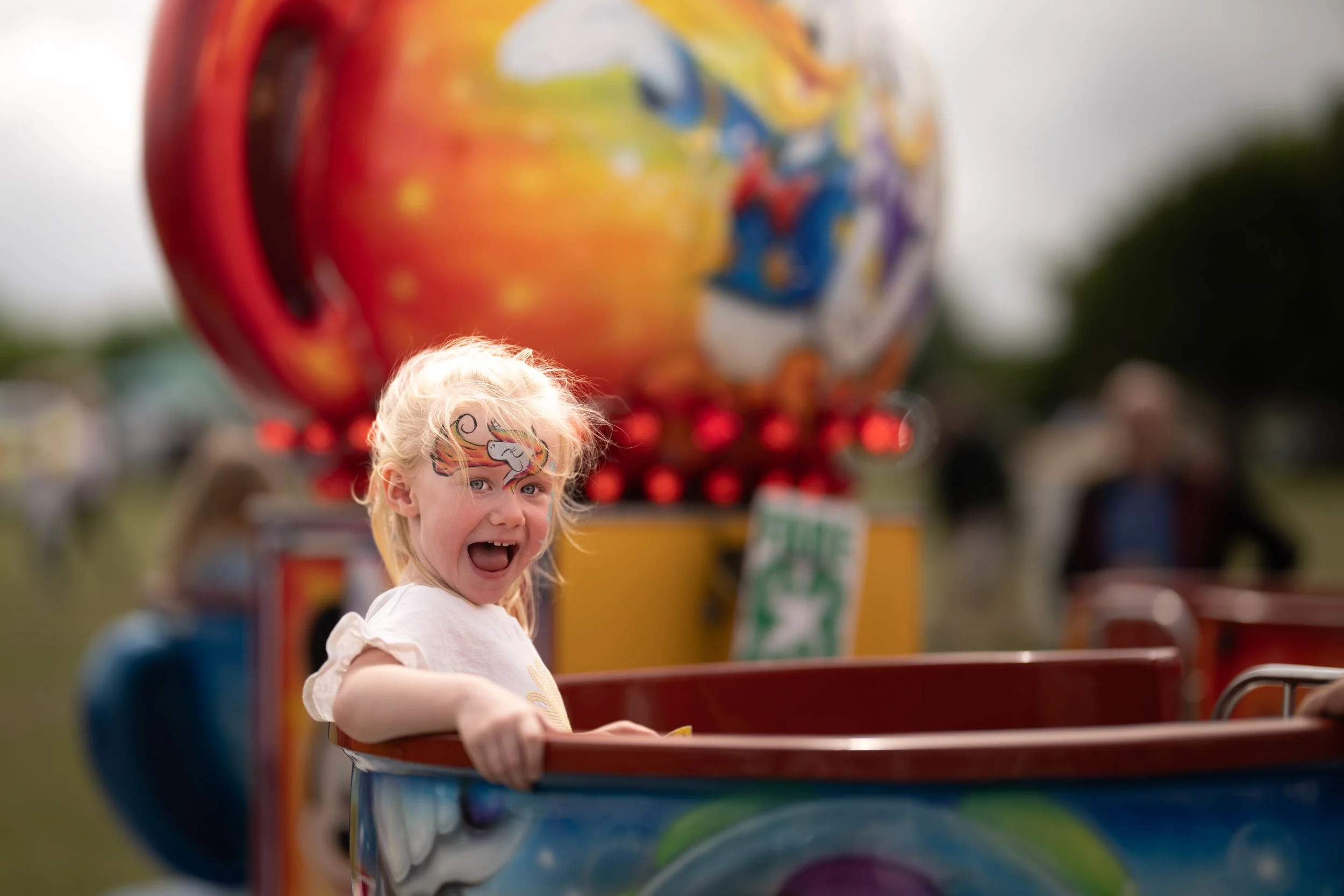 Young girl with face paint enjoying a carnival ride, with a large colorful carnival attraction in the background.