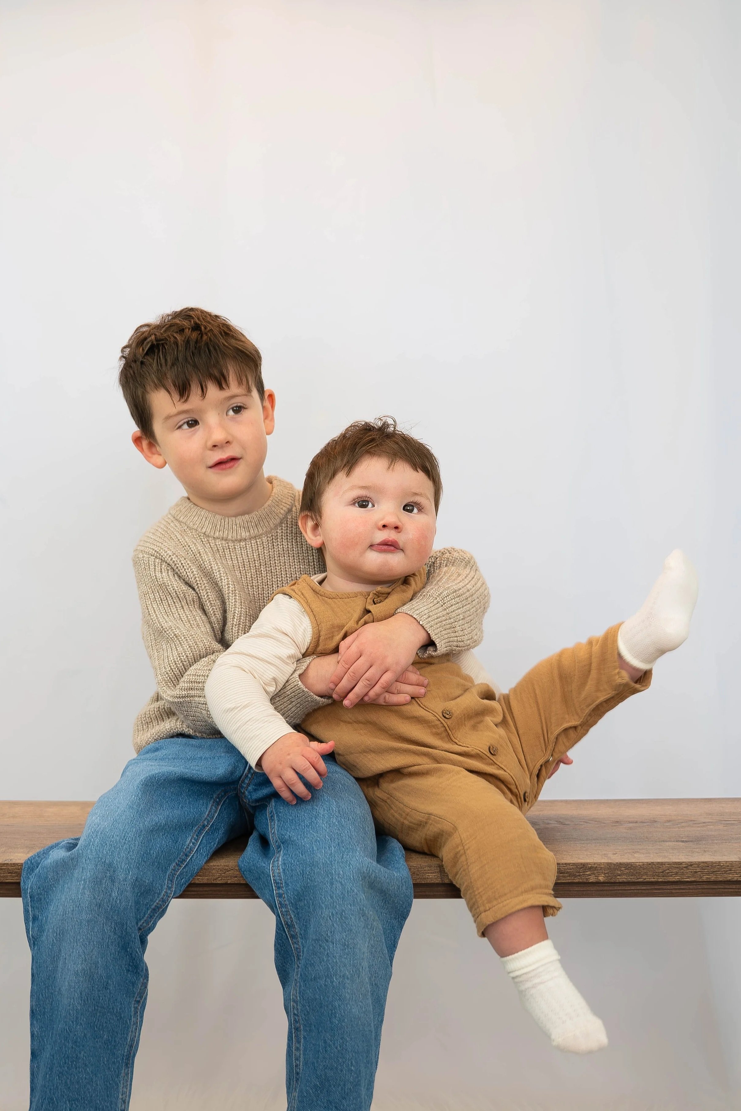 Two young boys, one older and one younger, sitting on a wooden bench against a plain white background. The older boy is holding the younger boy, who is extending one leg outward.