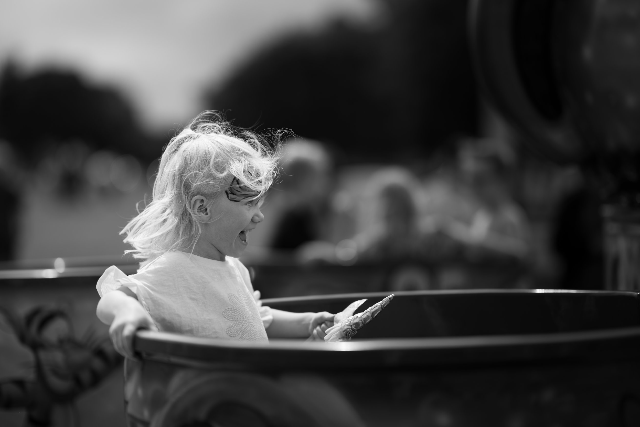 A young girl with light-colored hair and glasses, smiling and excited, sitting in a cart outdoors during daytime.