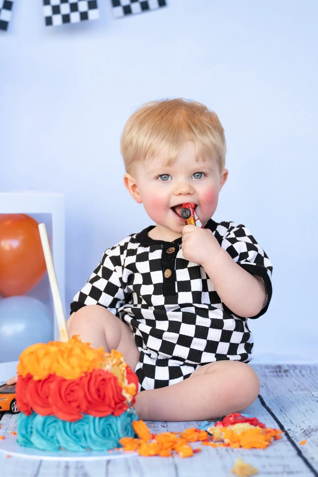 A young blond-haired boy in a black and white checkered shirt sitting on a floor with a colorful birthday cake in front of him. He is holding a small toy car in his mouth, surrounded by orange and red cake frosting and birthday decorations.