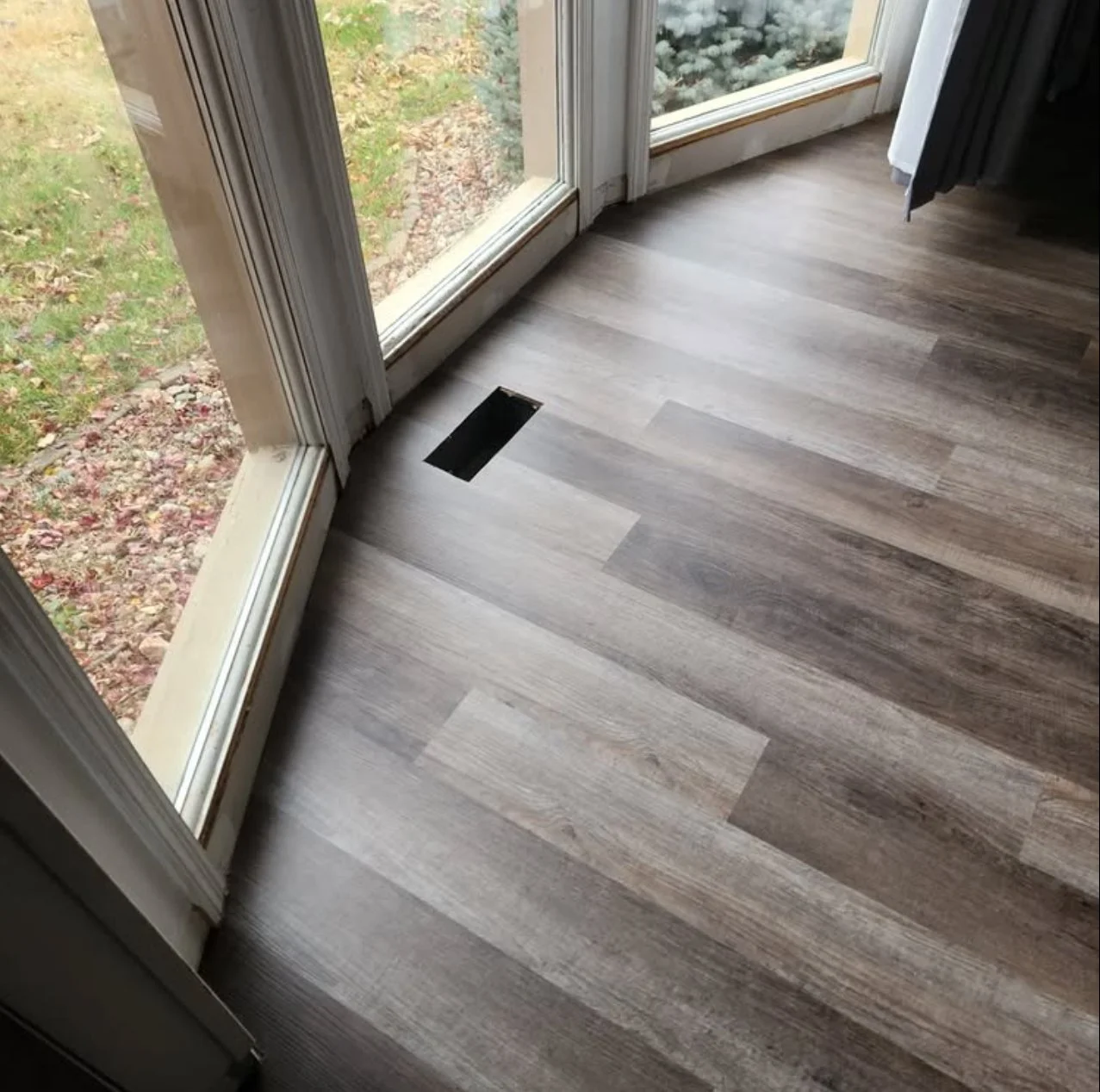 Interior view of a room with wood laminate flooring, adjacent to glass sliding doors leading outside to a yard with grass and fallen leaves. There is an air vent on the floor.