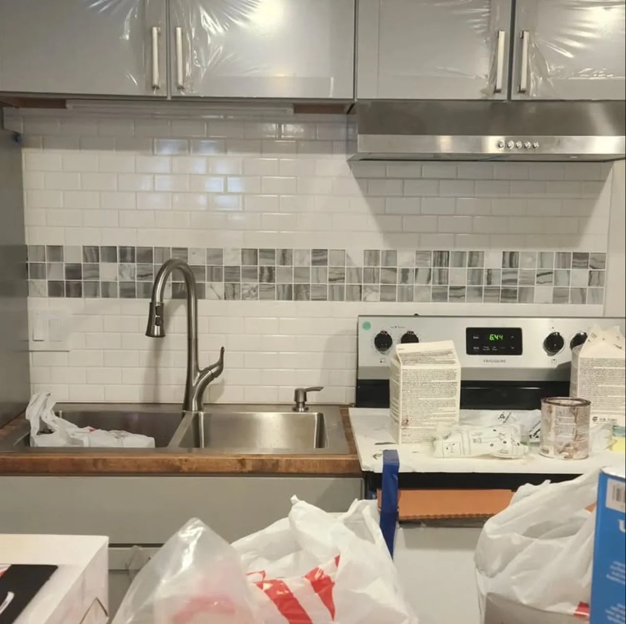 A cluttered kitchen with a stainless steel sink, a modern faucet, a white countertop, a white tiled backsplash with gray accent tiles, and a white stove with black controls, surrounded by various items and packaging.