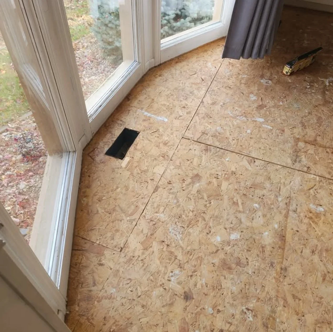 Close-up of a new plywood floor with an air vent near the glass door, and a partially opened curtain in a room.