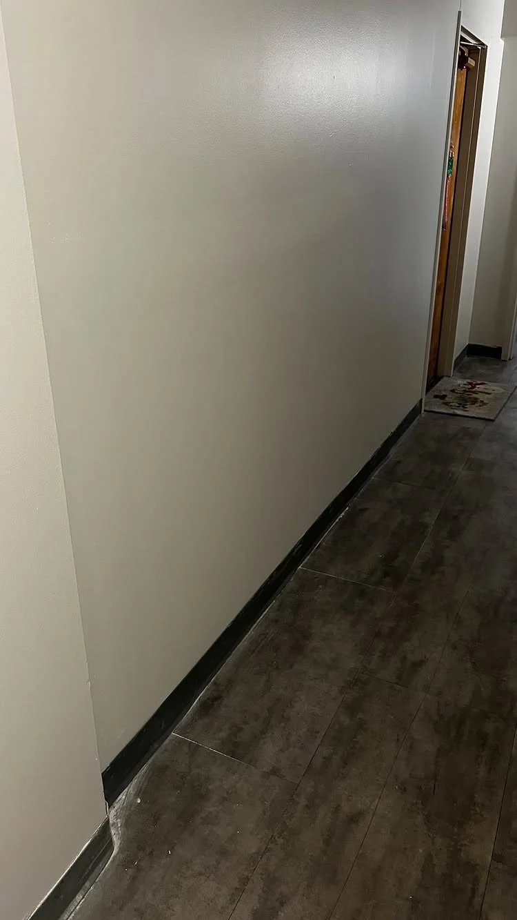 Photo of a plain off-white wall and a brown tiled floor in an indoor space, with a partially visible doorway on the right side.