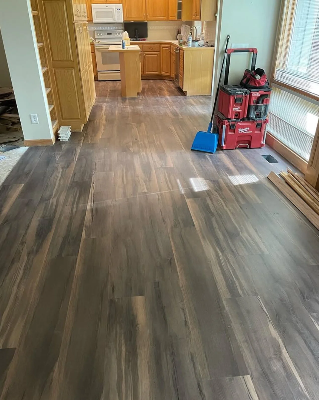 Interior of a kitchen with hardwood flooring being installed. Tools and equipment, including a red toolbox, a dustpan, and wooden planks, are visible near a large window.