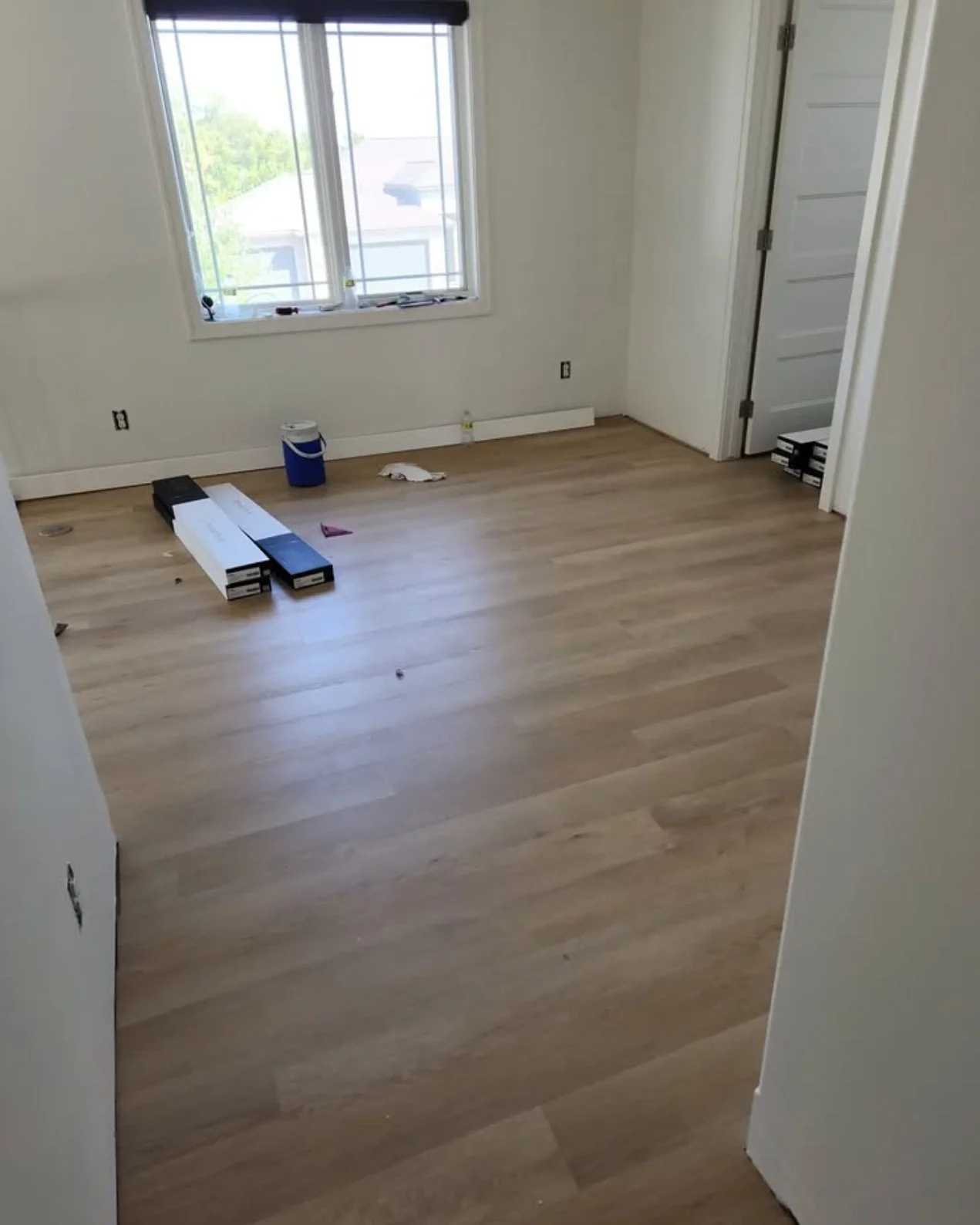 Empty room with light-colored hardwood floors, a window with a view outside, and various construction tools and materials on the floor.