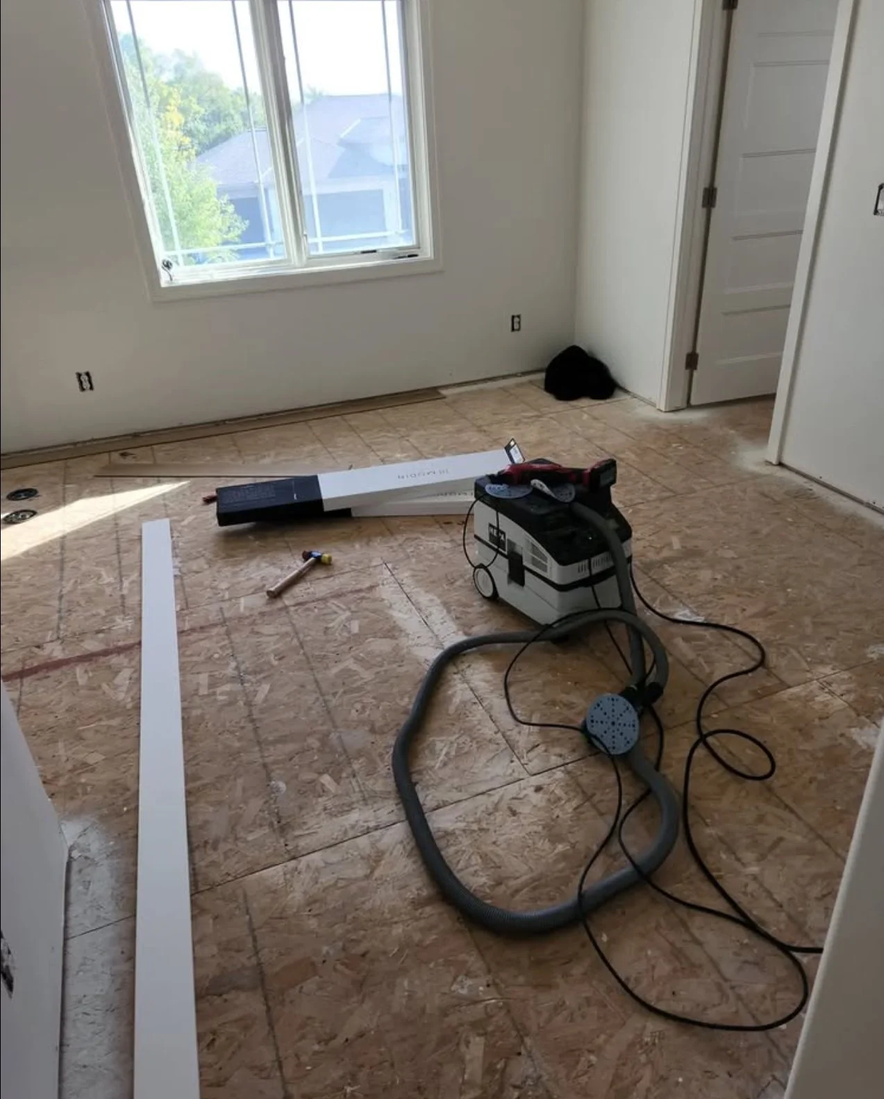 Room with a large window, construction tools, vacuum, and a black object in the corner of the room.