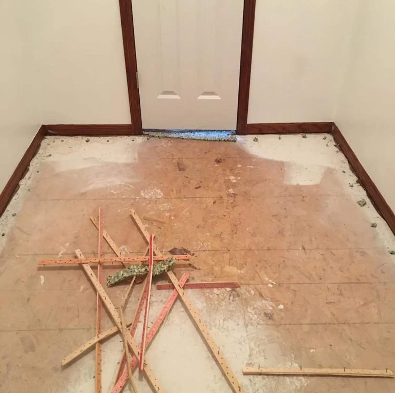 Room with a partially removed wooden floor, construction tools and materials on the floor, closed white door with brown trim.