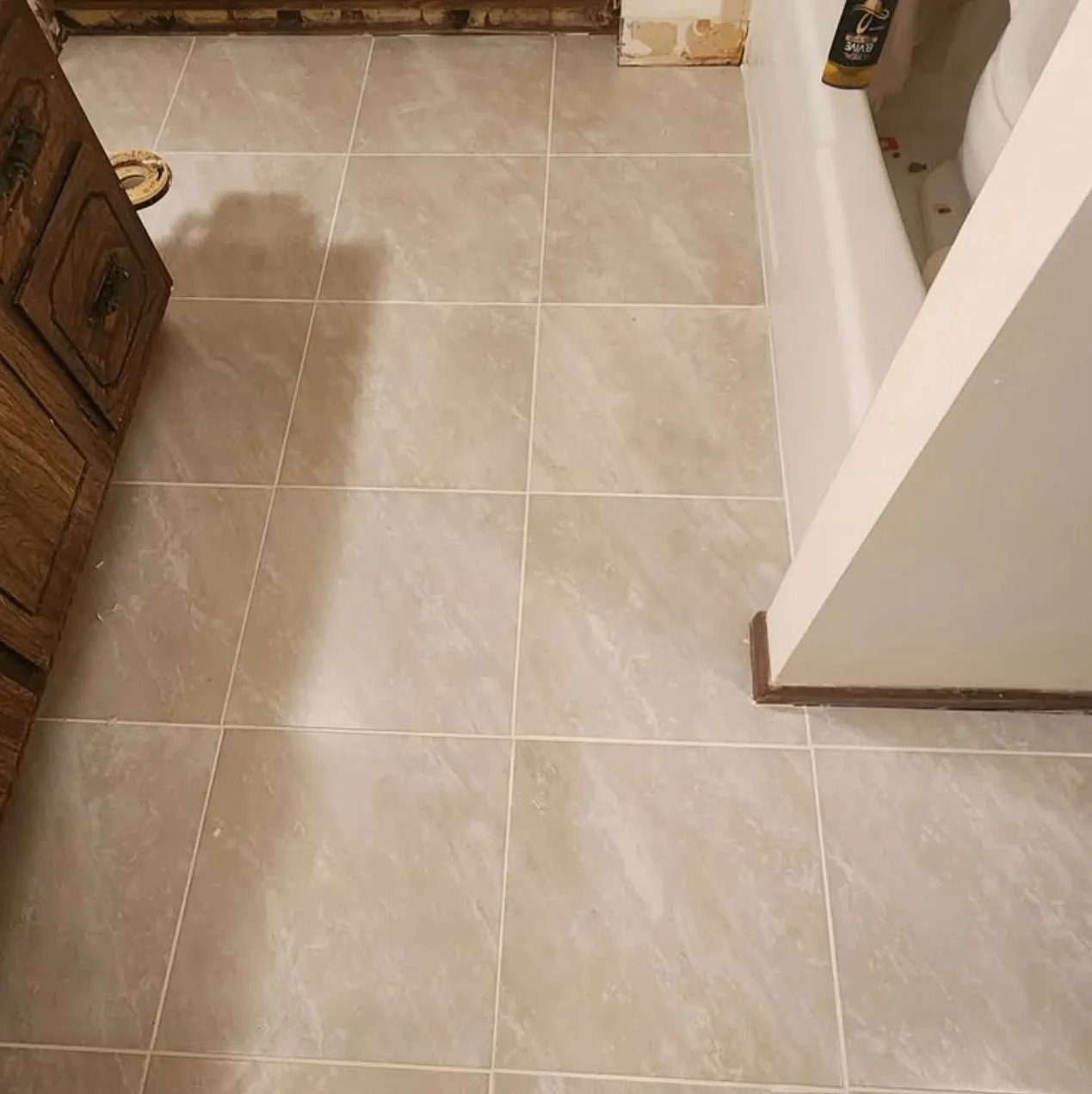 A bathroom floor with beige ceramic tiles, part of a wooden cabinet on the left, and a white bathtub on the right. A bottle of cleaning product is on the edge of the bathtub.