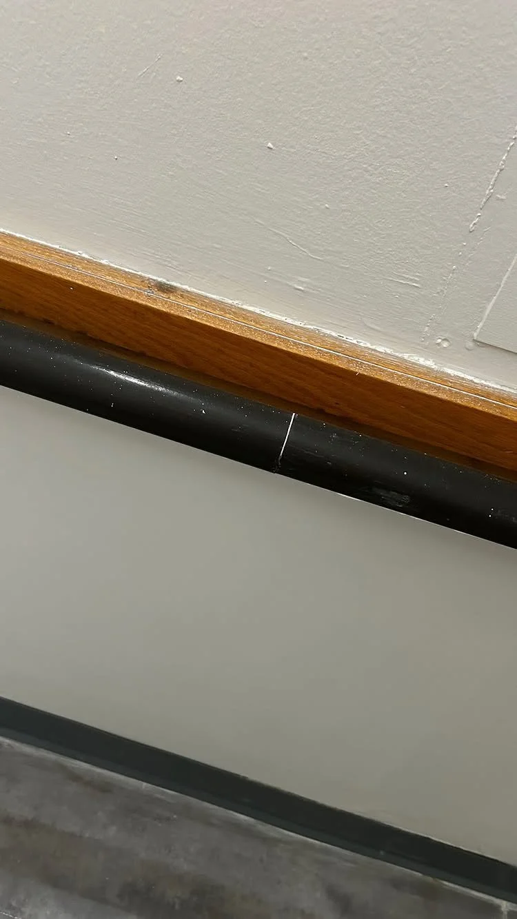 Close-up of a wall with a crack and a black pipe running along the baseboard, with a wooden trim at the top.