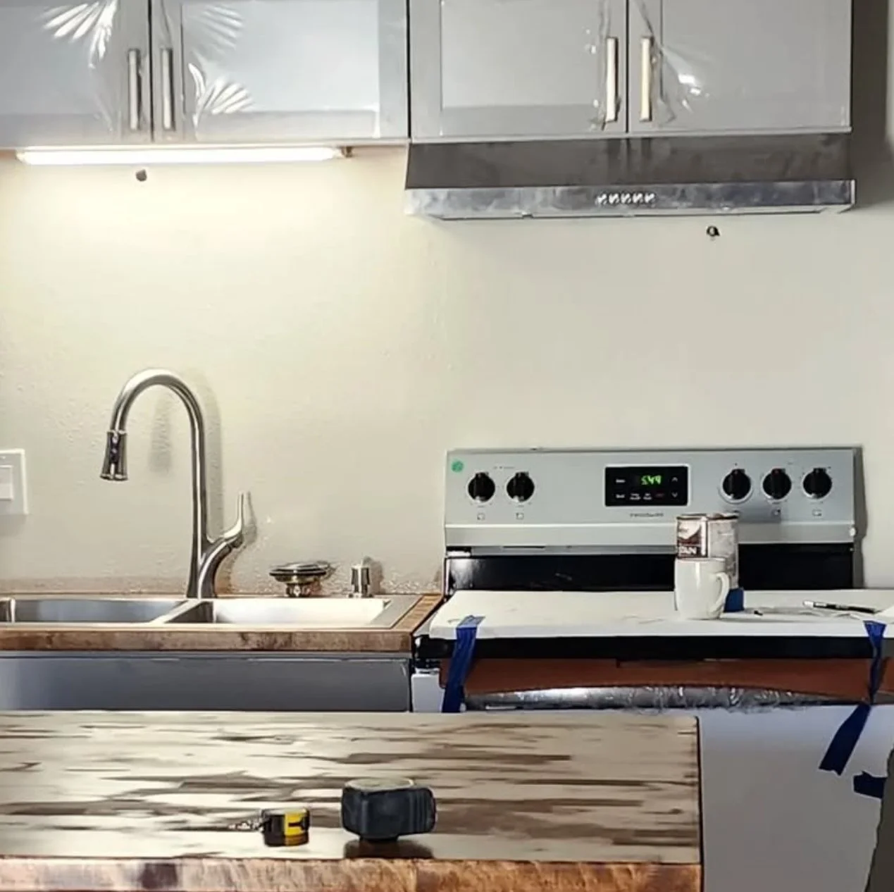 Kitchen with stainless steel upper cabinets, a double sink with a tall curved faucet, a stovetop with a digital display, a mug, and a wooden countertop with tools on it, indicating ongoing renovation.