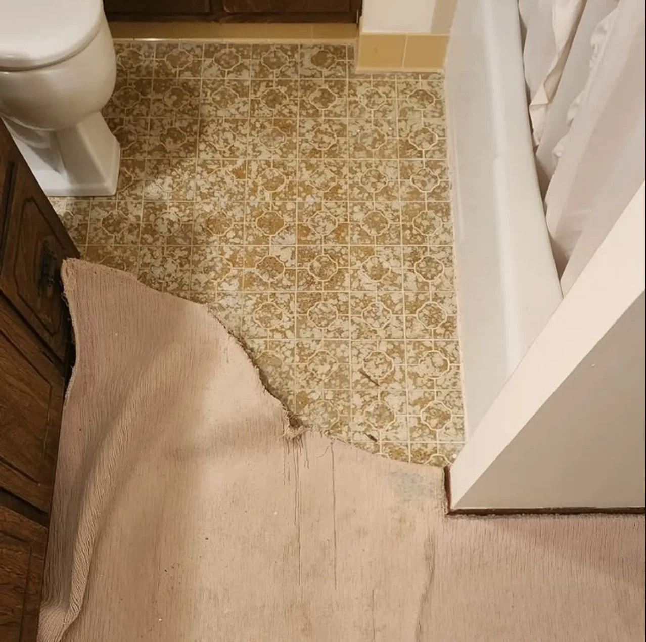 A bathroom corner showing a damaged beige carpet peeling back to reveal old patterned linoleum flooring underneath. The toilet and part of a wooden cabinet are visible.