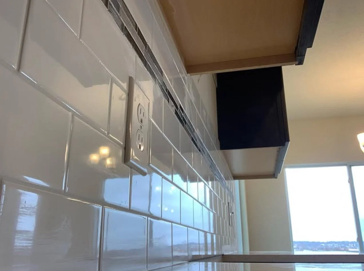Close-up of a kitchen countertop with white subway tiles, an electrical outlet, and a beige and brown kitchen cabinet. There is a window in the background showing a cloudy sky and outdoor landscape.