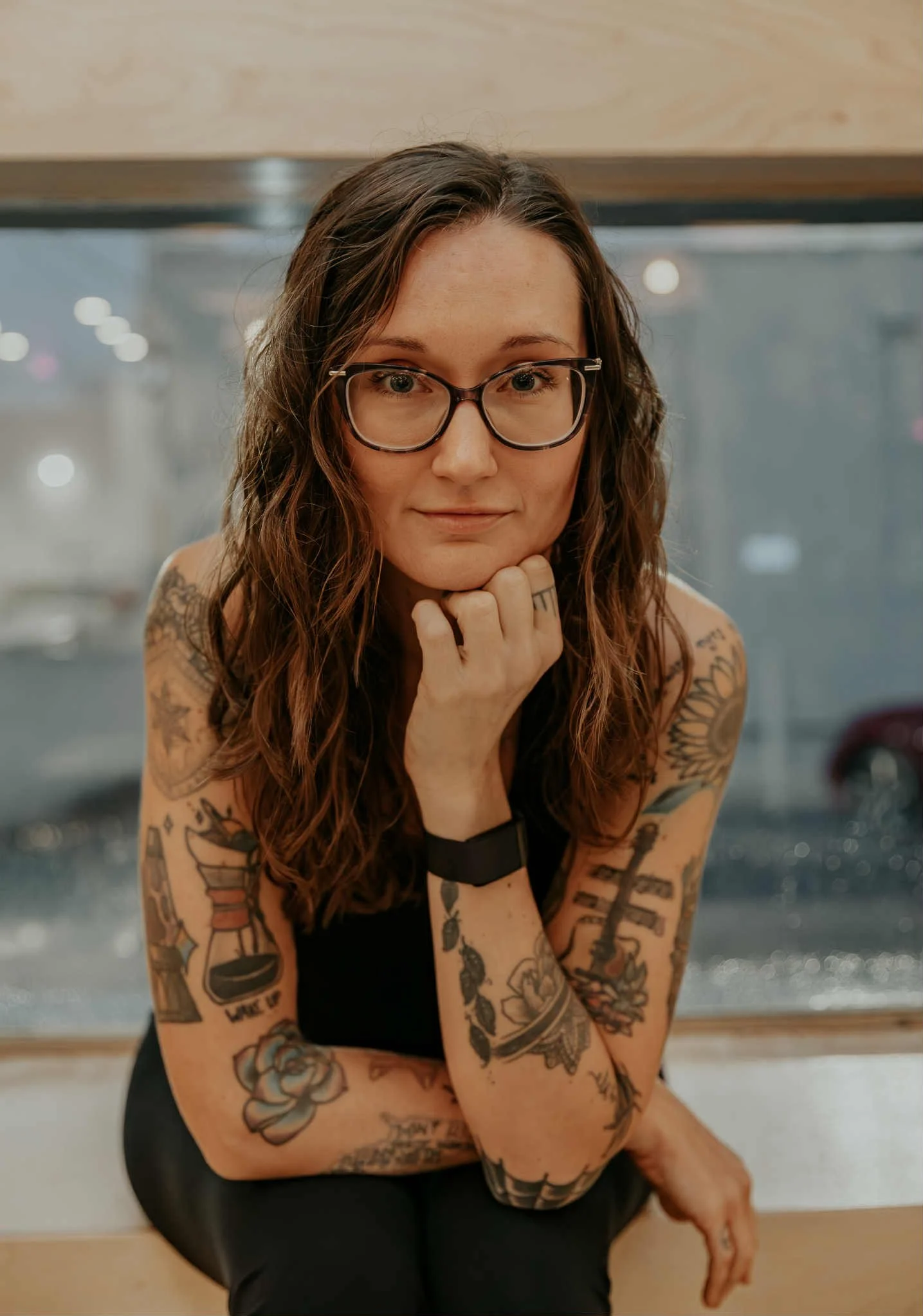 Portrait of a somatic wellness coach with wavy brown hair and glasses, sitting in a reflective pose with tattooed arms. Calm, approachable expression suitable for a holistic healing and mind-body wellness website. 