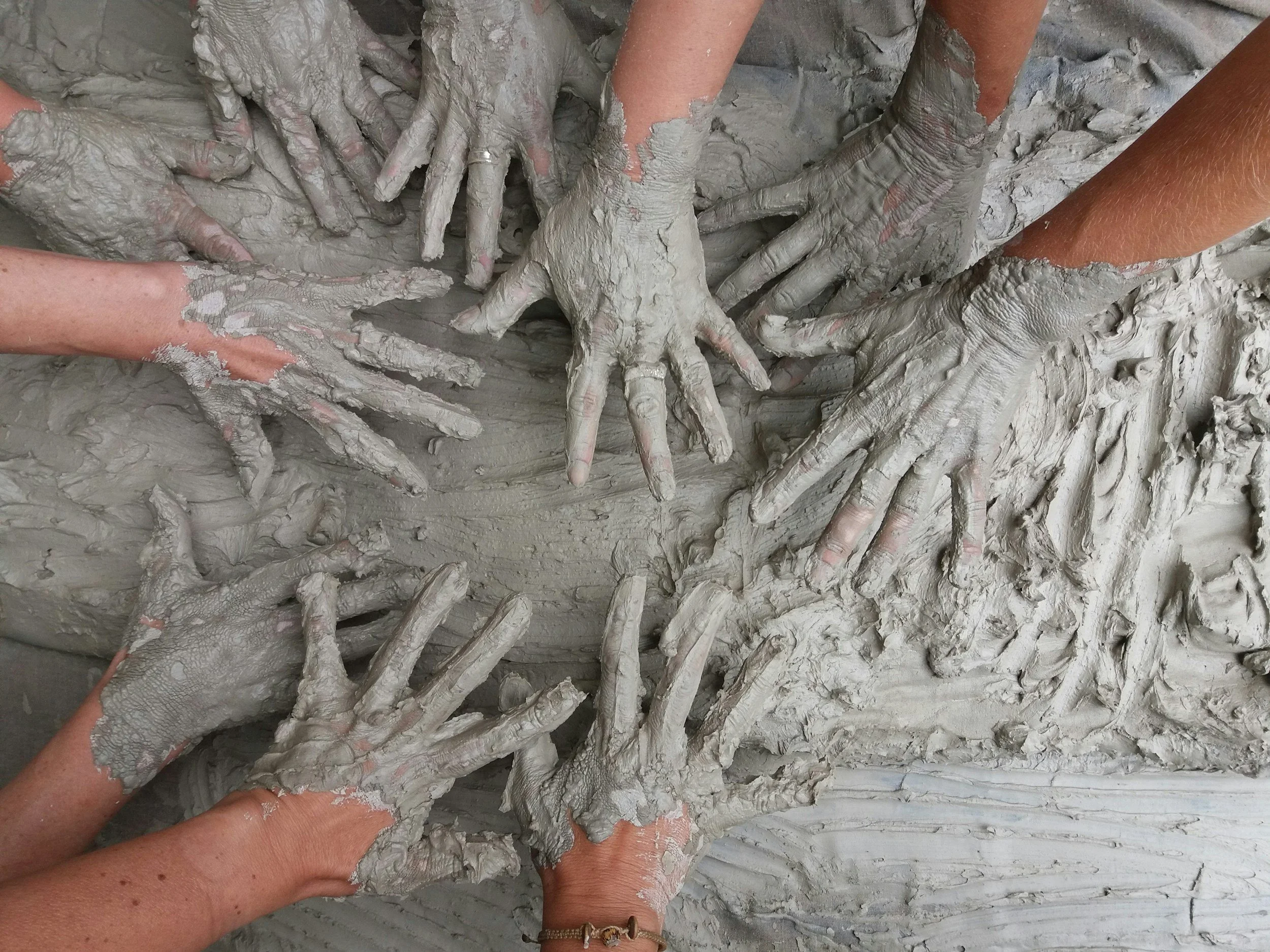 Multiple hands covered in wet gray mud, working together to mold or shape the mud.