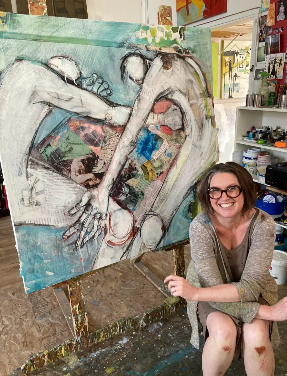 Artist seated beside a large mixed-media figurative artwork. The image reflects themes of embodiment, emotional expression, creativity, and storytelling through visual art, and is well suited for pages focused on art,creative process, healing. 