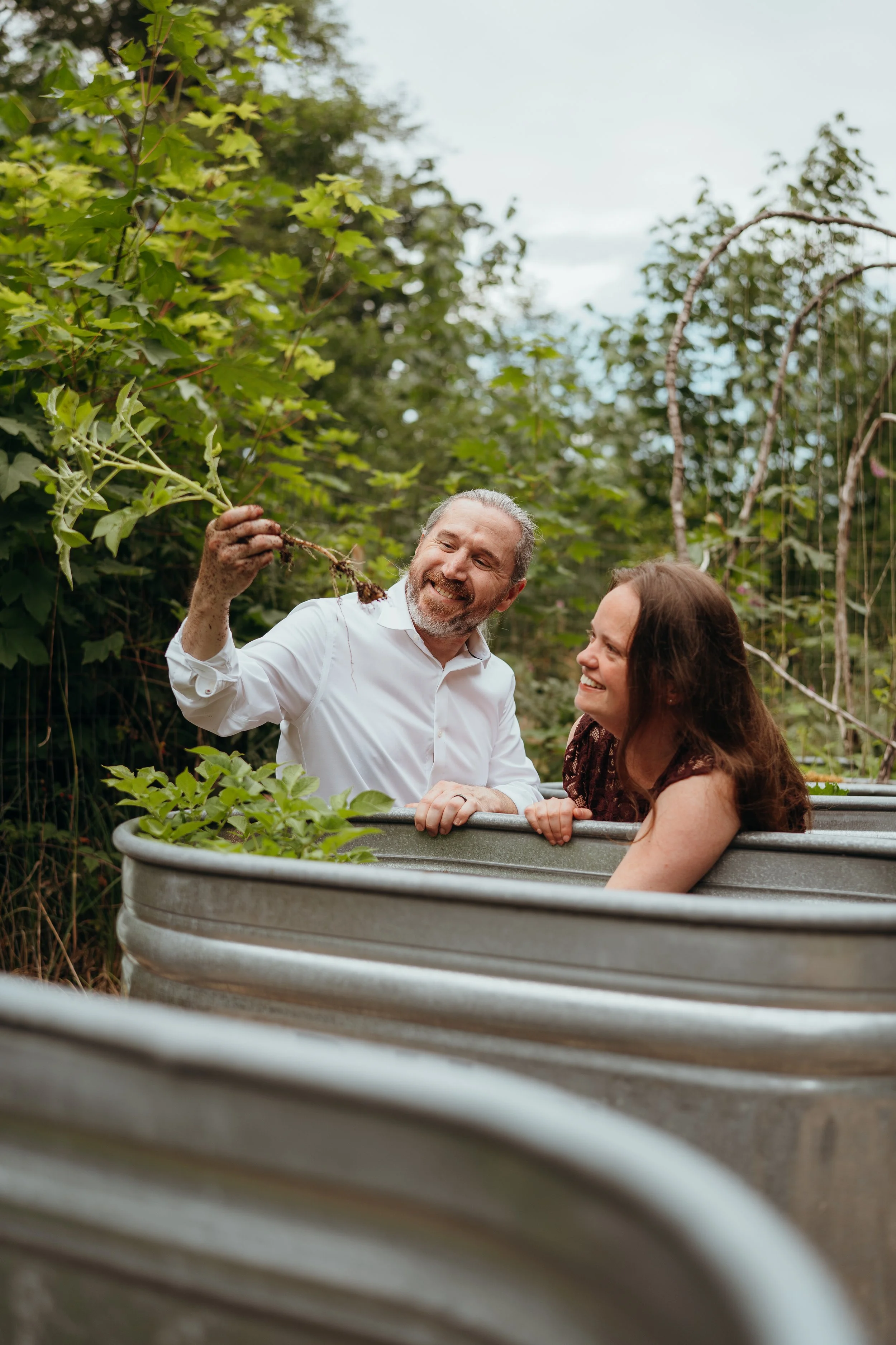 A man and woman smiling and talking while standing in a metal tub filled with soil and plants, surrounded by green trees in a garden or outdoor setting.