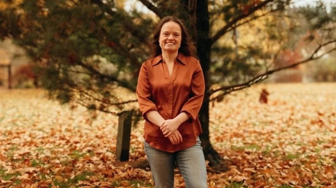 A woman standing outdoors in fall foliage, smiling, wearing an orange shirt and jeans.