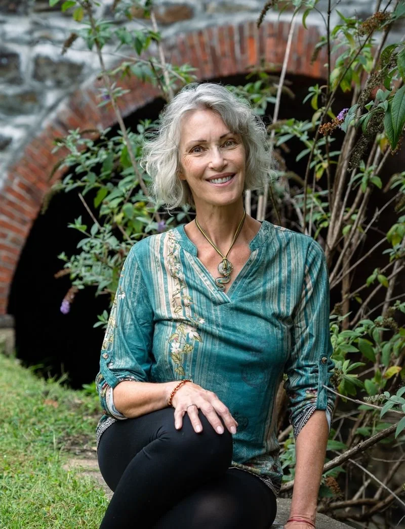 A smiling older woman with gray, curly hair sitting on grass in front of a brick archway and green plants.