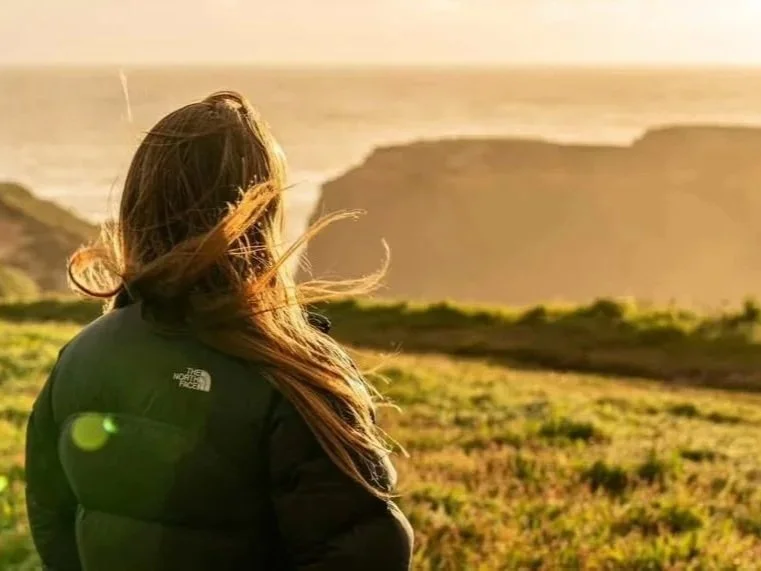 A woman with long hair standing outdoors, looking at a sunset or sunrise over a hilly landscape near the ocean.