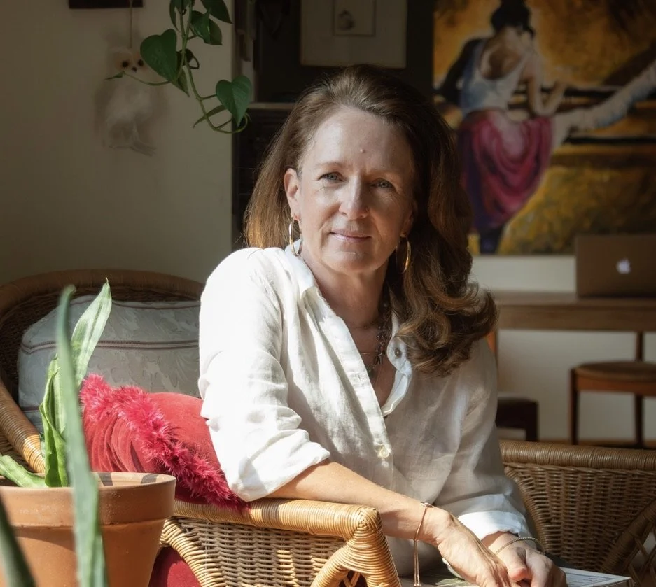 A woman with long brown hair, wearing a white shirt and large hoop earrings, sitting on a wicker chair with a red pillow and green plants around her in a sunlit room, with a colorful painting and a laptop in the background.