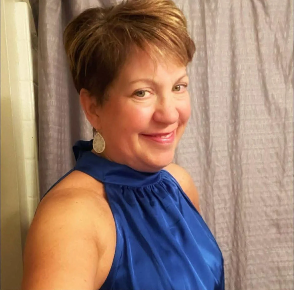 A middle-aged woman with short brown hair smiling, wearing a royal blue sleeveless dress and gold earrings, standing in front of a light-colored curtain.