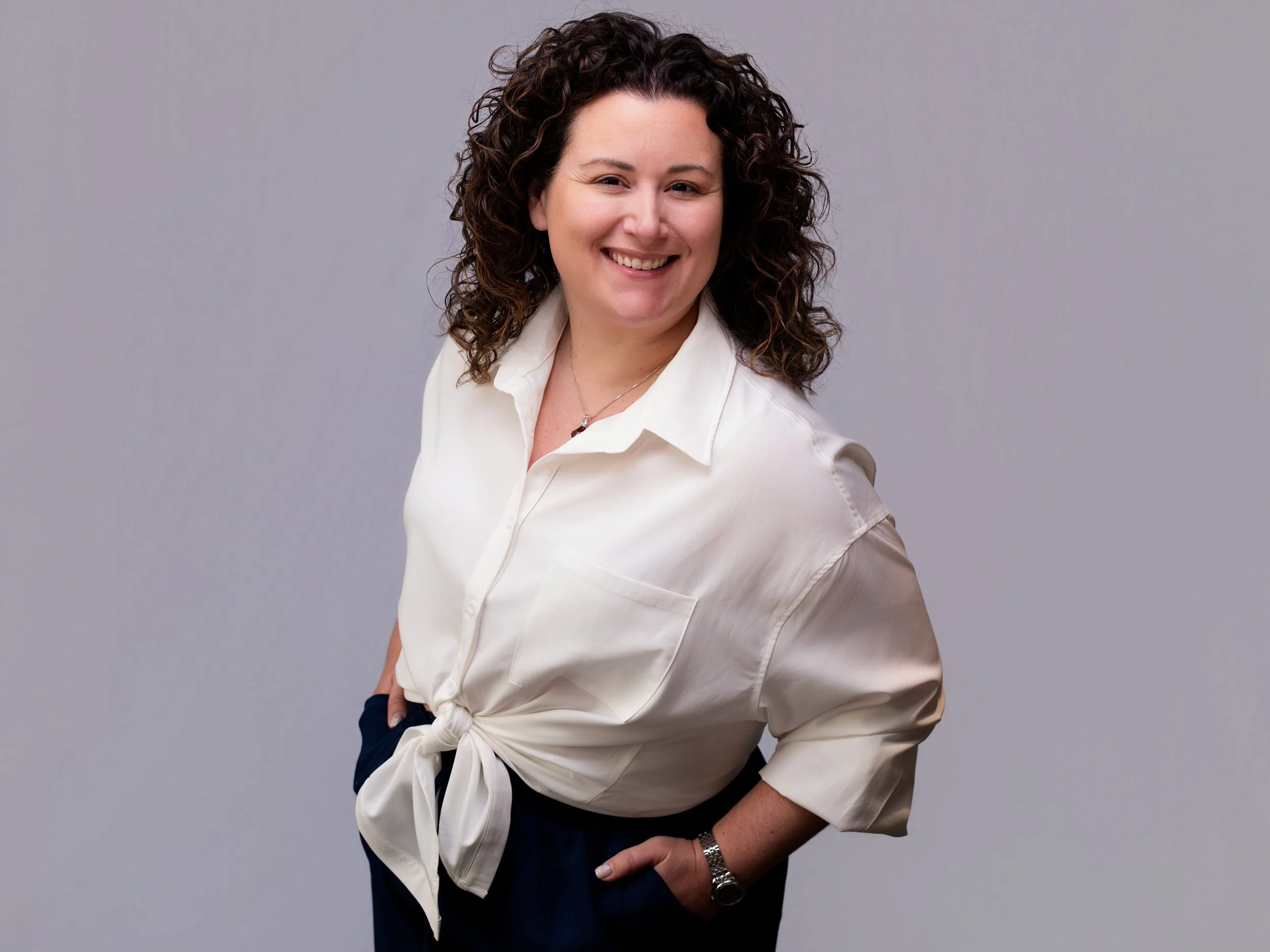 A woman with curly hair wearing a white shirt tied at the waist, smiling, against a plain gray background.
