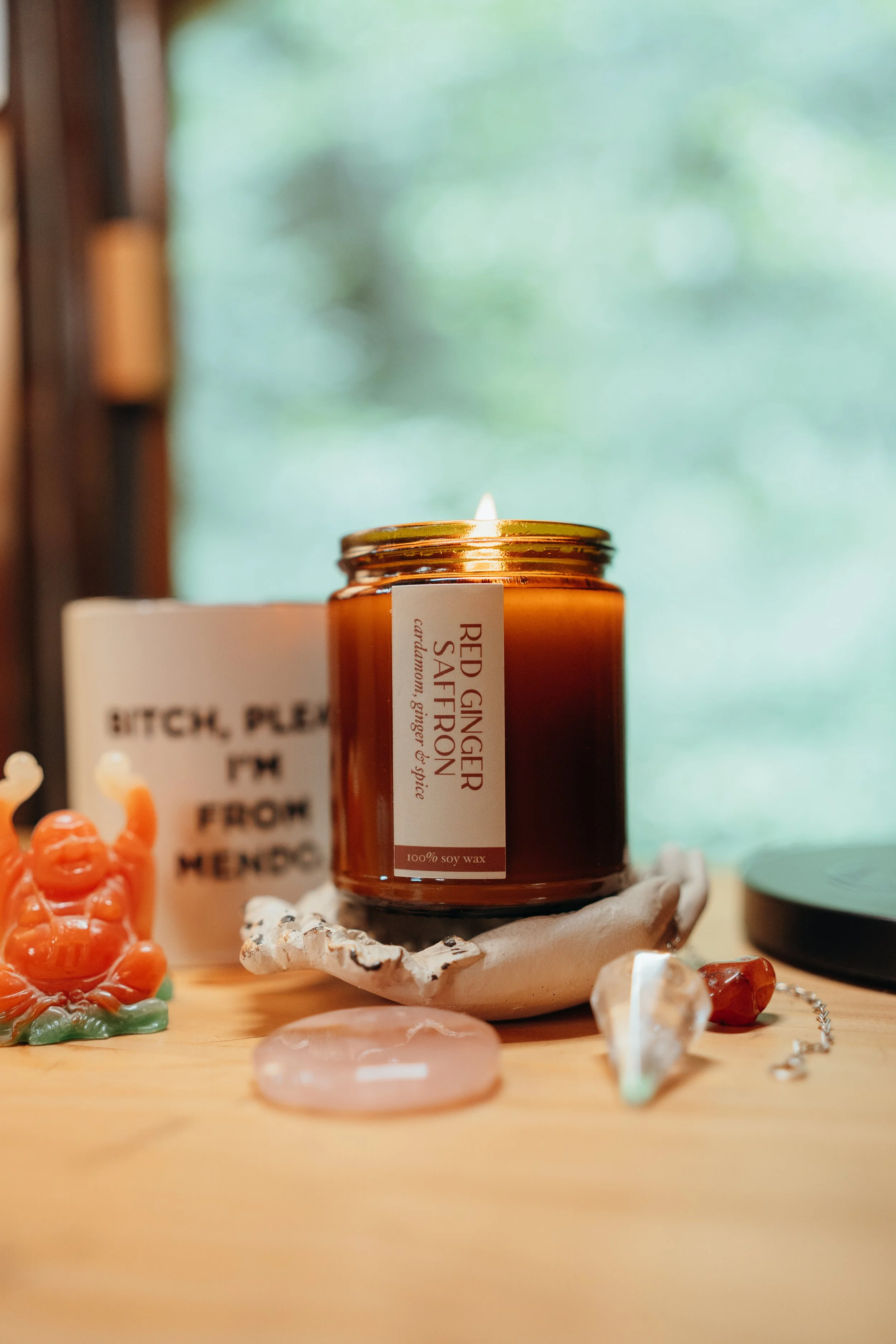 A candle in a brown jar labeled 'Red Ginger Saffron,' placed on a seashell. Surrounding it are various small decorative objects including a orange figurine, a pink stone, and jewelry, with a blurred background of a window with green trees outside.
