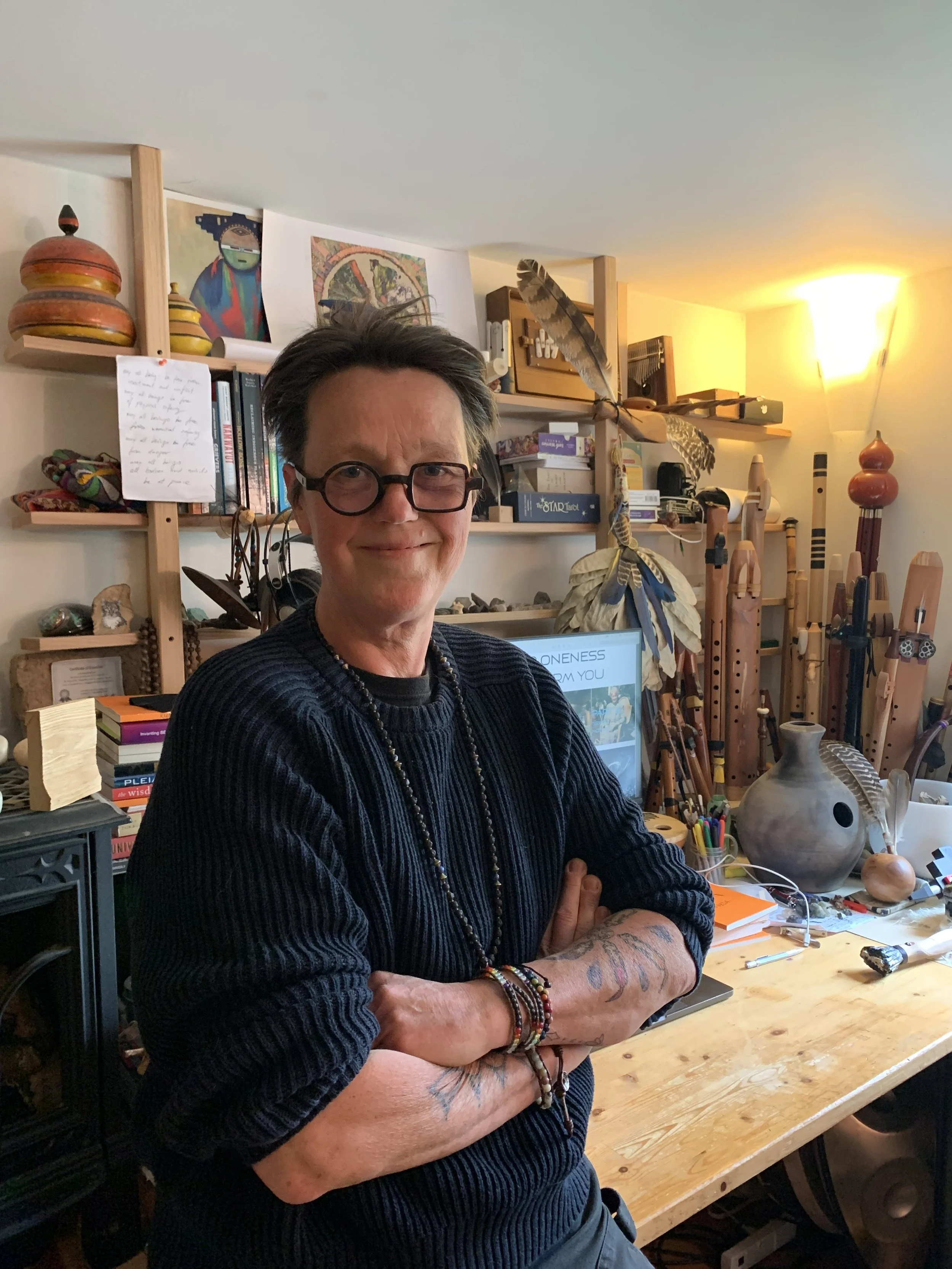 A man with glasses and tattoos on his arms standing with arms crossed in a cluttered room filled with books, art, and musical instruments, with artwork and a feather decoration on the shelves behind him, and a laptop on a wooden table.