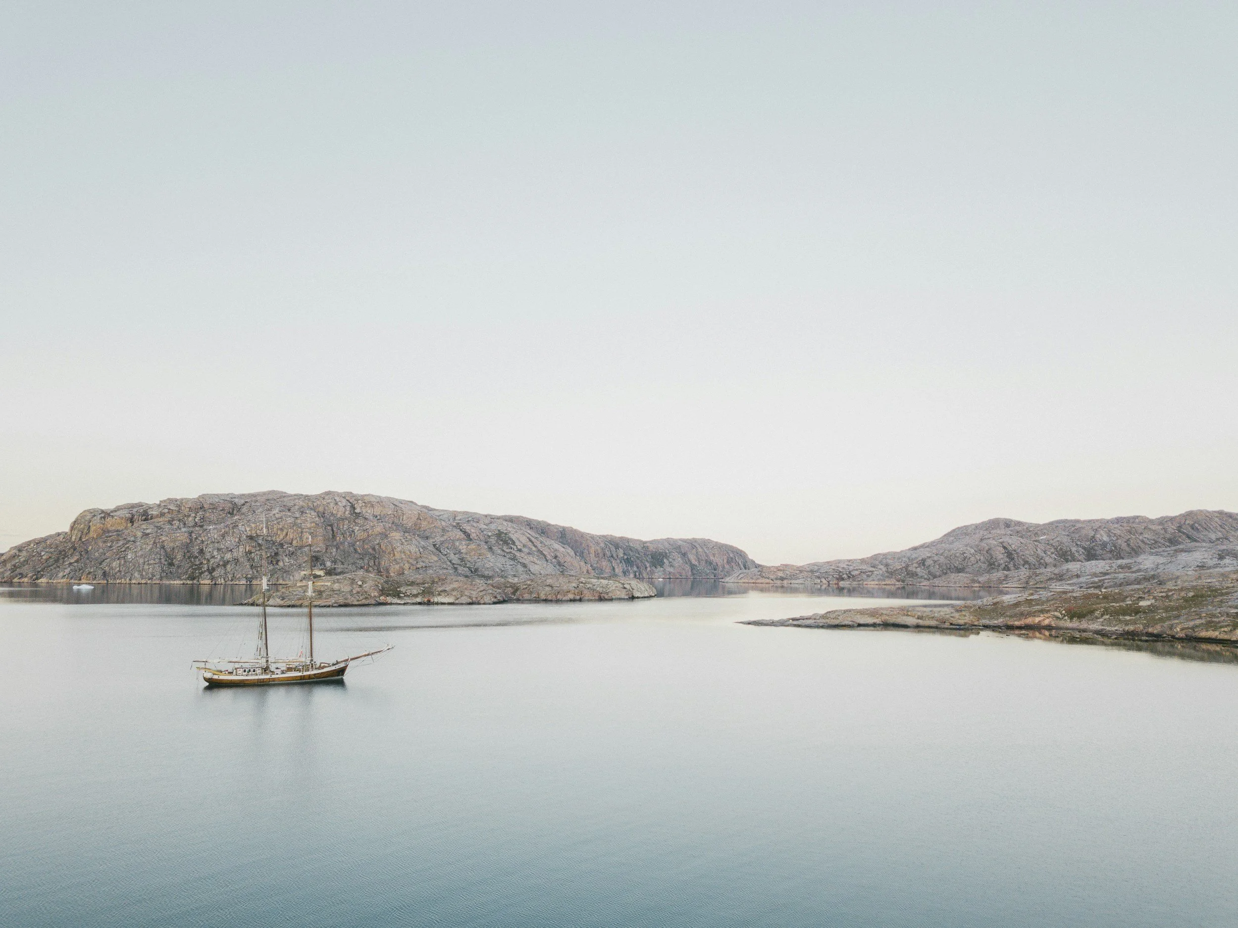 A sailboat floating on calm water near rocky islands under a pale sky.