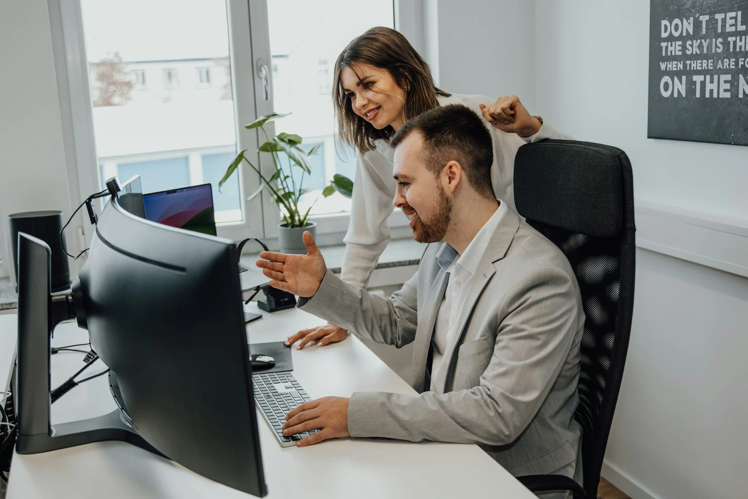 Two colleagues, a woman and a man, are having a discussion at a computer in an office. The woman is standing and smiling, while the man is seated and gesturing towards the screen.