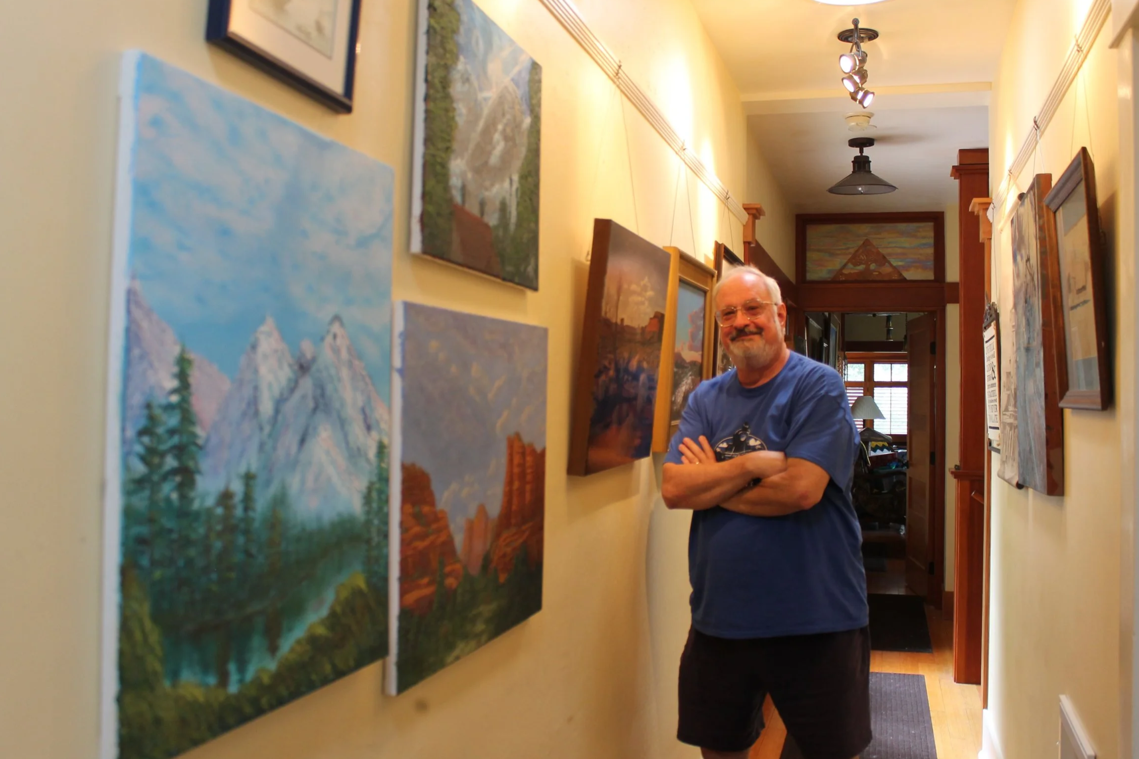 Photo of artist Tom Selinske standing in a hallway lined with his landscape paintings of mountains, forests, and red rock canyons