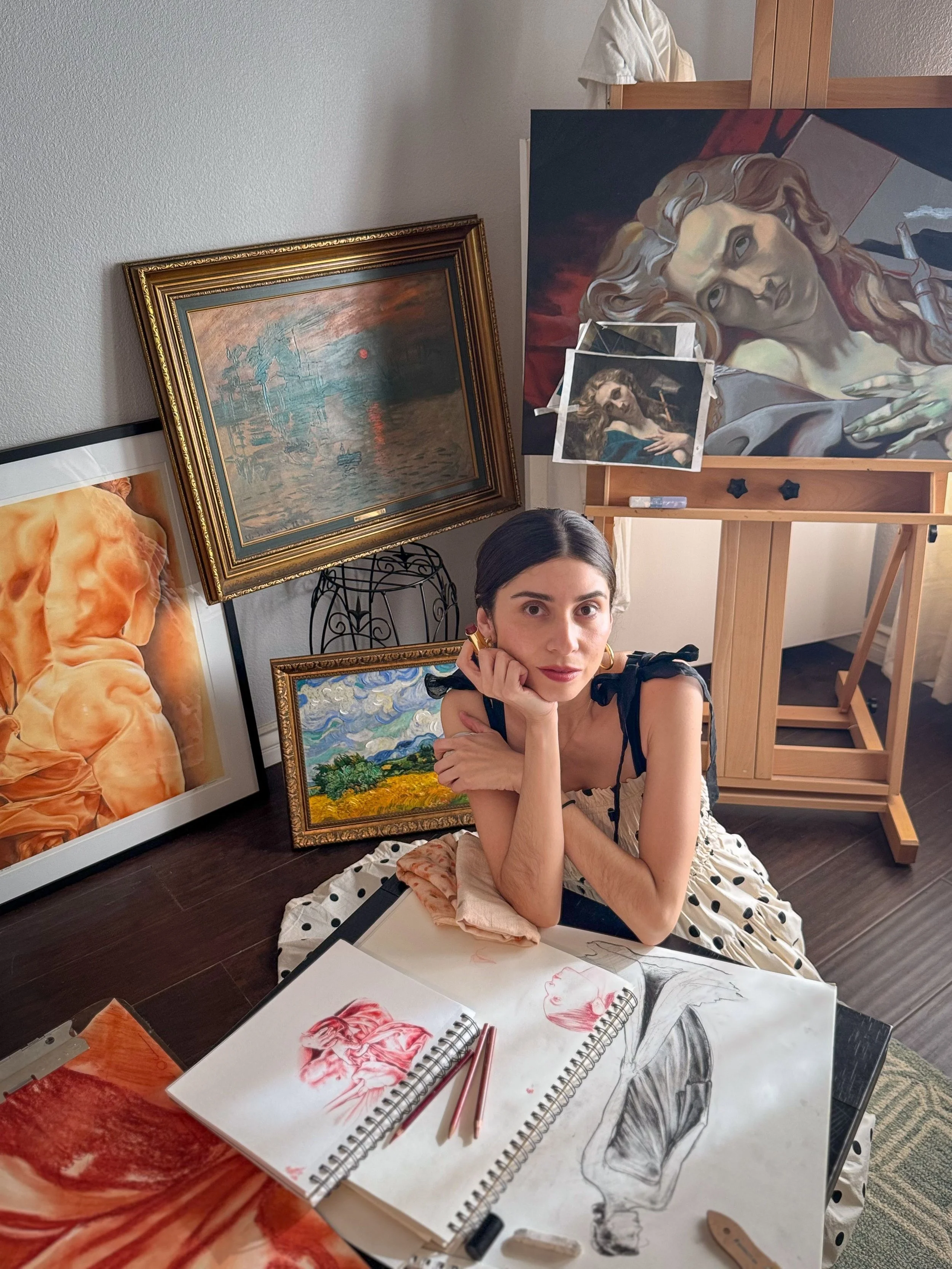 Photo of artist Bianca Ornelas in her studio, seated at a table with sketches, surrounded by paintings including a seaside landscape, a wheat field, and a study of a reclining sculptural figure