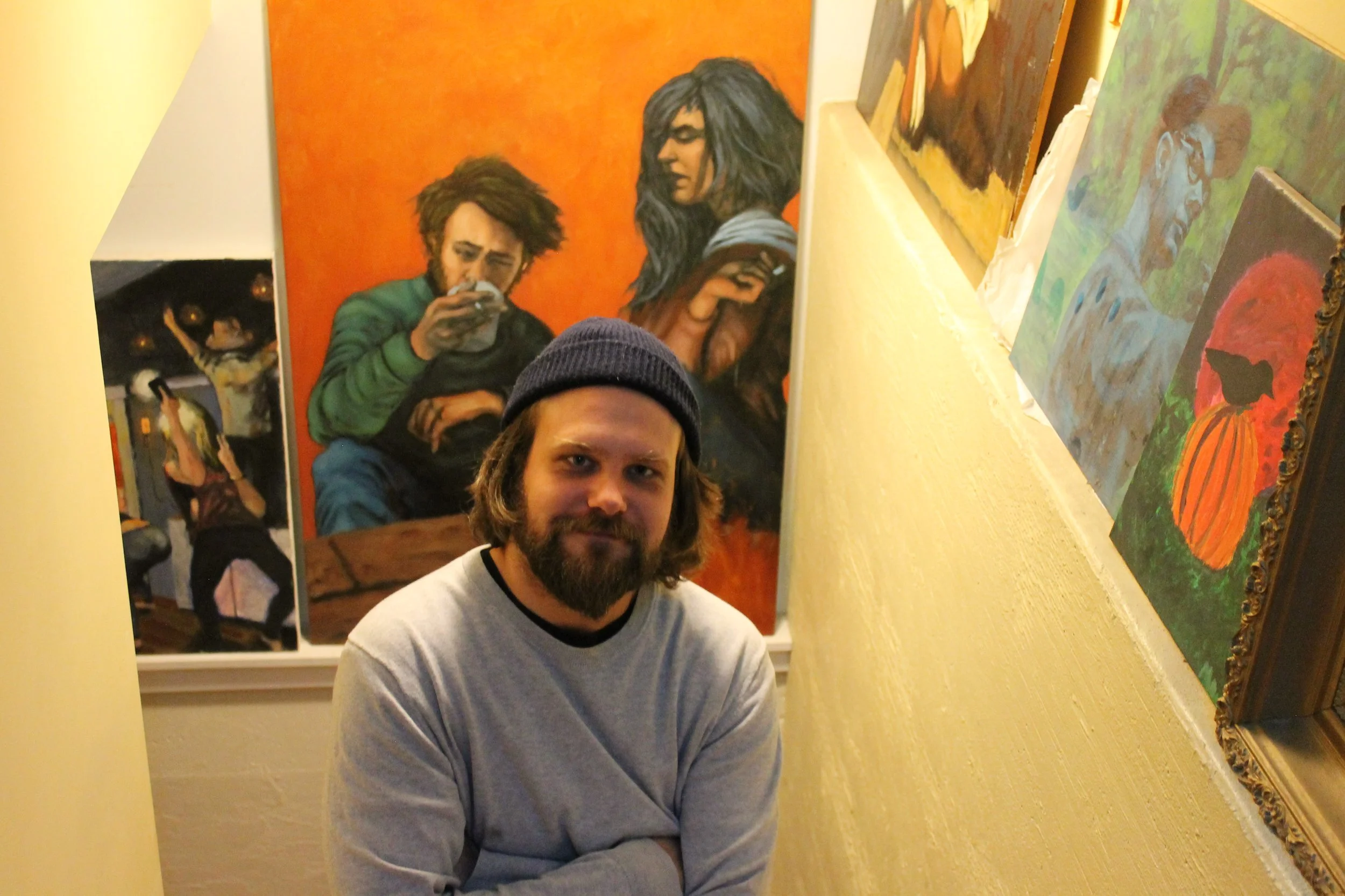 Photo of artist Paul Selinske, wearing a beanie and crew-neck sweater, seated on a staircase surrounded by several of his paintings, including an orange-toned portrait of two figures