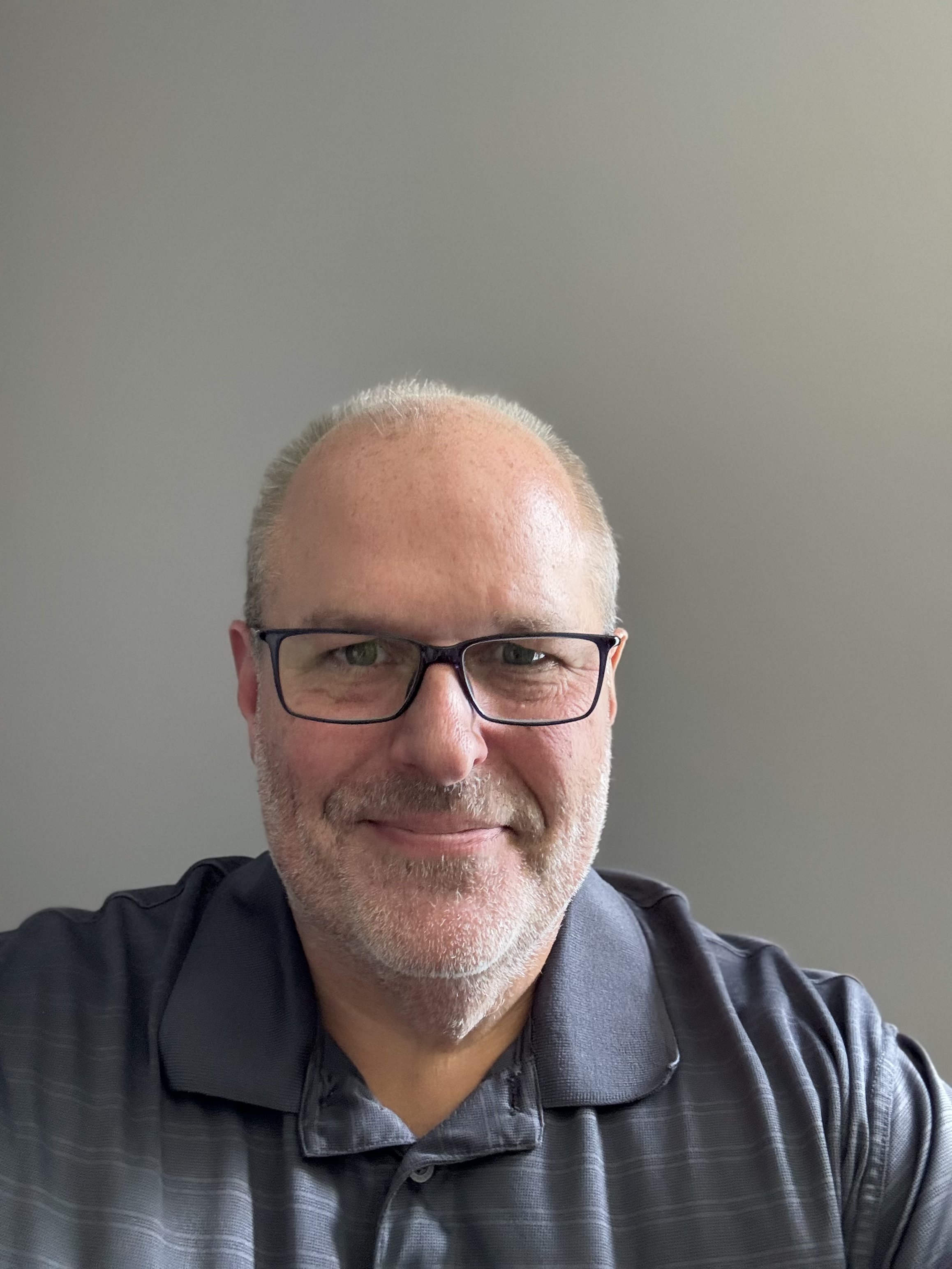 A middle-aged man with glasses and a short gray beard, wearing a dark gray polo shirt, taking a selfie in front of a plain gray wall.