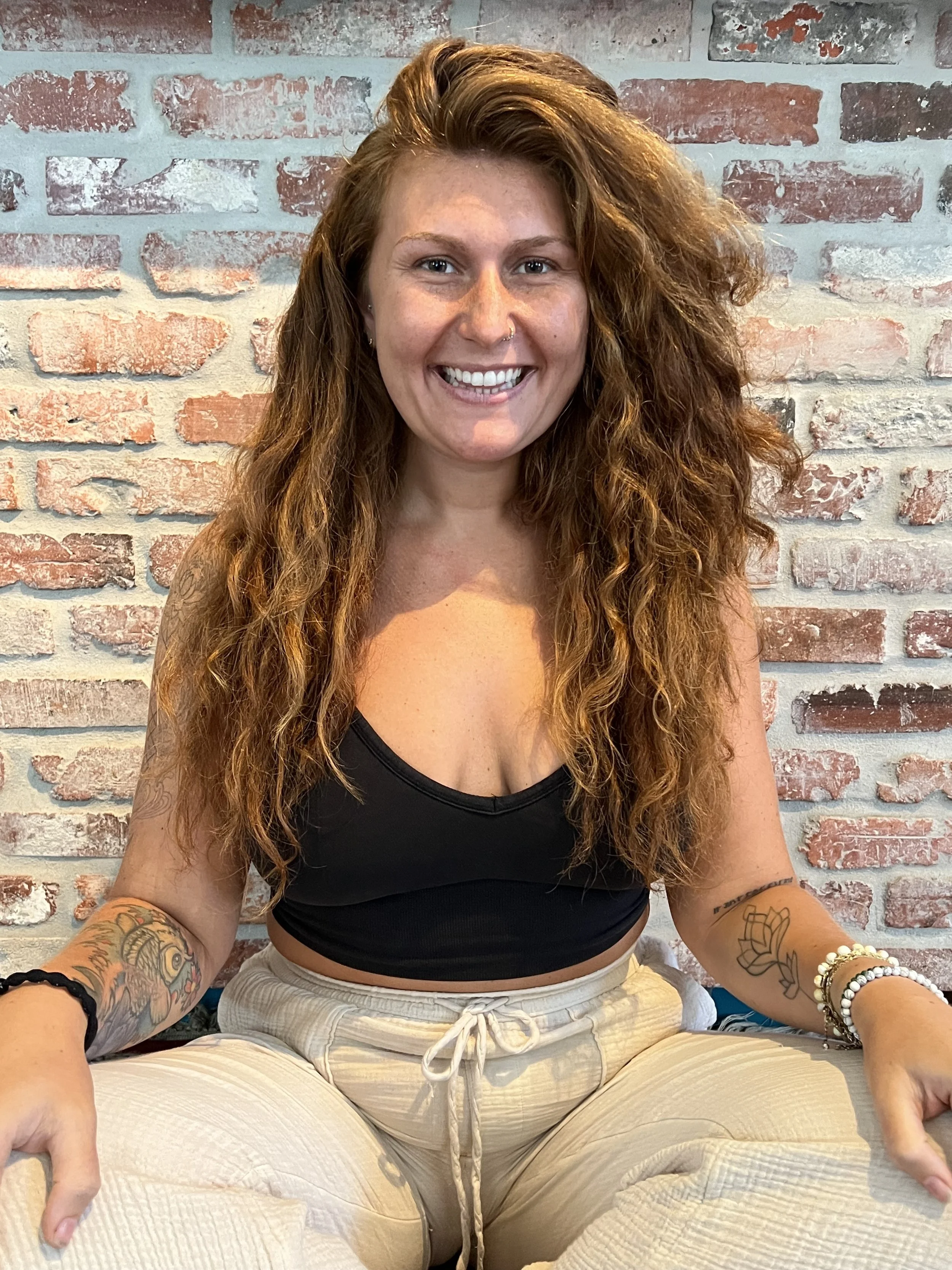 A woman with long curly red hair smiling while sitting cross-legged against a brick wall. She has tattoos on her arms, wears bracelets, and a nose ring, and is dressed in a black top and beige pants.